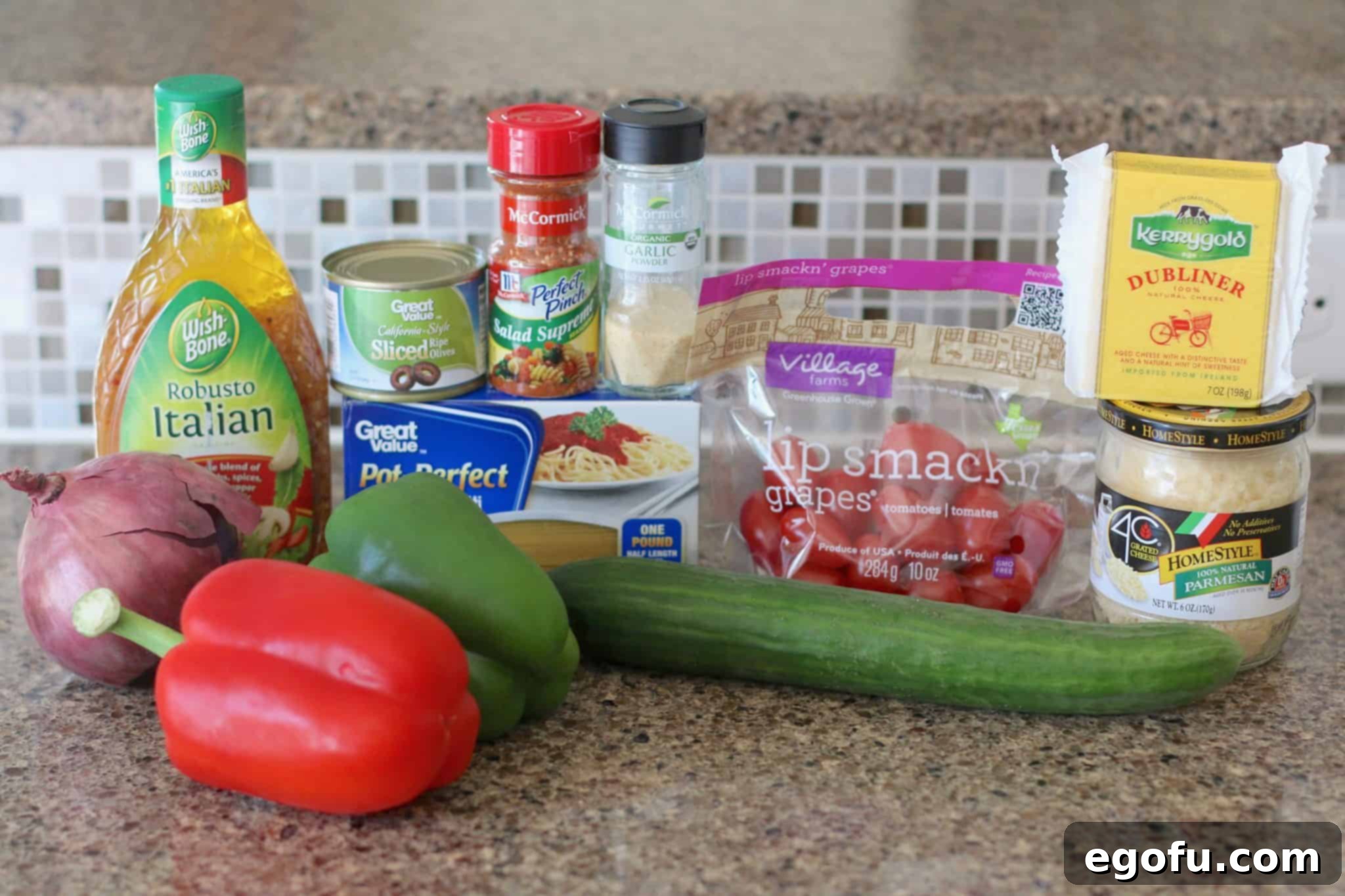 A flat lay photograph showcasing all the fresh ingredients for Summer Spaghetti Salad: spaghetti noodles, green and red bell peppers, red onion, English cucumber, grape tomatoes, sliced olives, cheddar cheese, grated Parmesan cheese, McCormick Salad Supreme seasoning, garlic powder, and Wish-Bone Italian dressing.