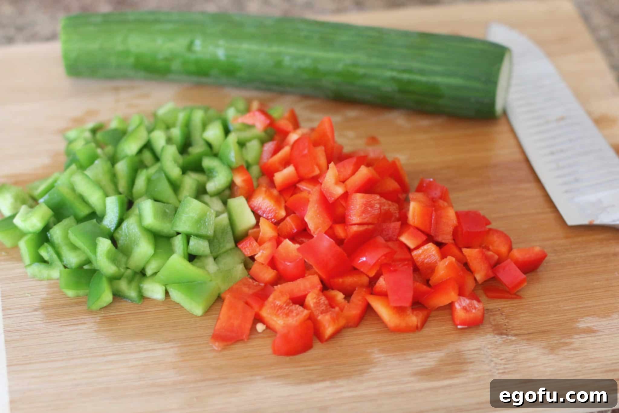 Freshly diced cucumbers, green peppers, and red peppers neatly arranged on a wooden cutting board, prepped for the salad.