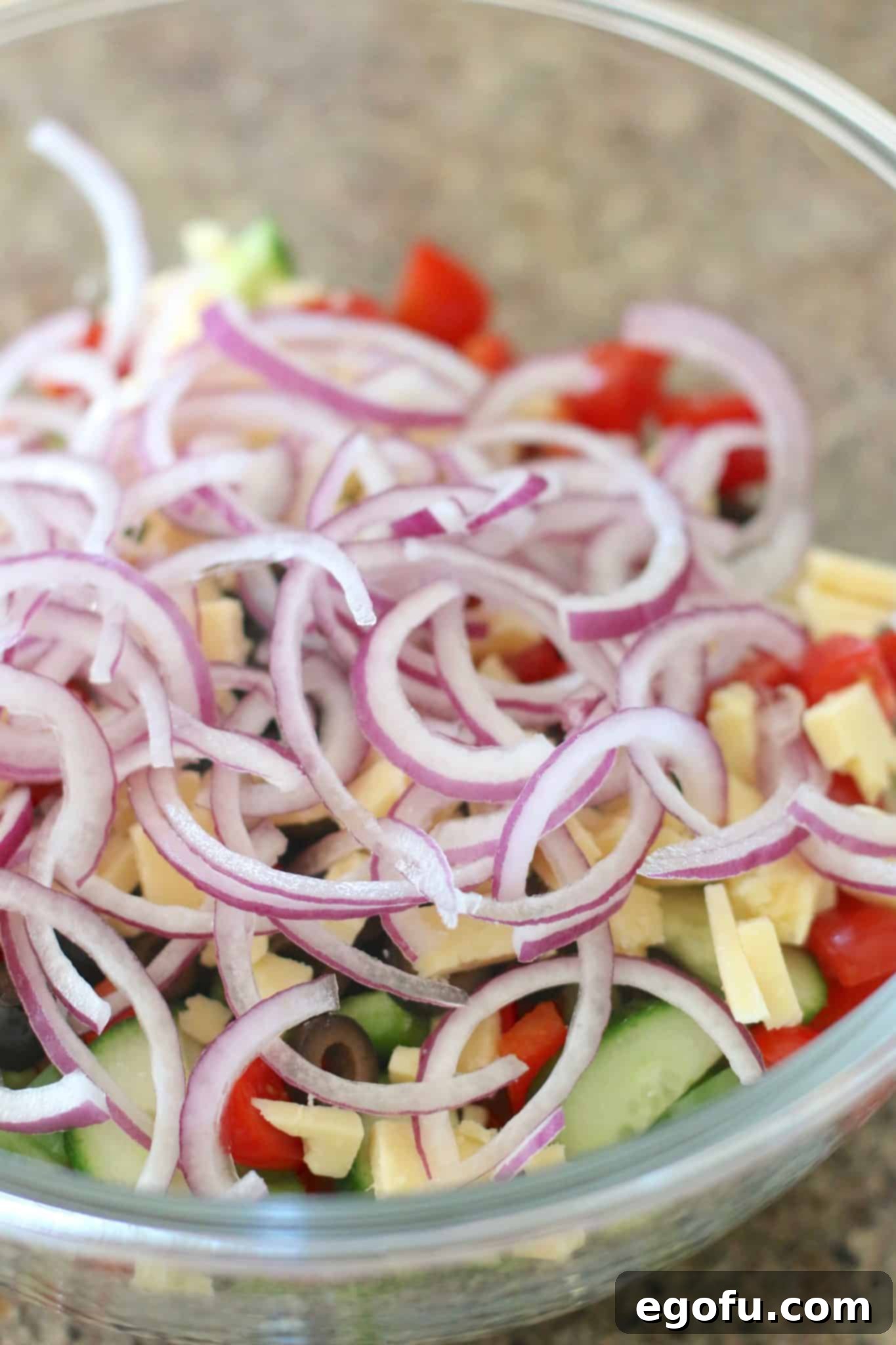 A vibrant mix of cooked spaghetti noodles, diced cheese, sliced red onion, cherry tomatoes, green and red bell peppers, cucumber, and sliced olives in a large clear glass bowl, ready to be tossed.