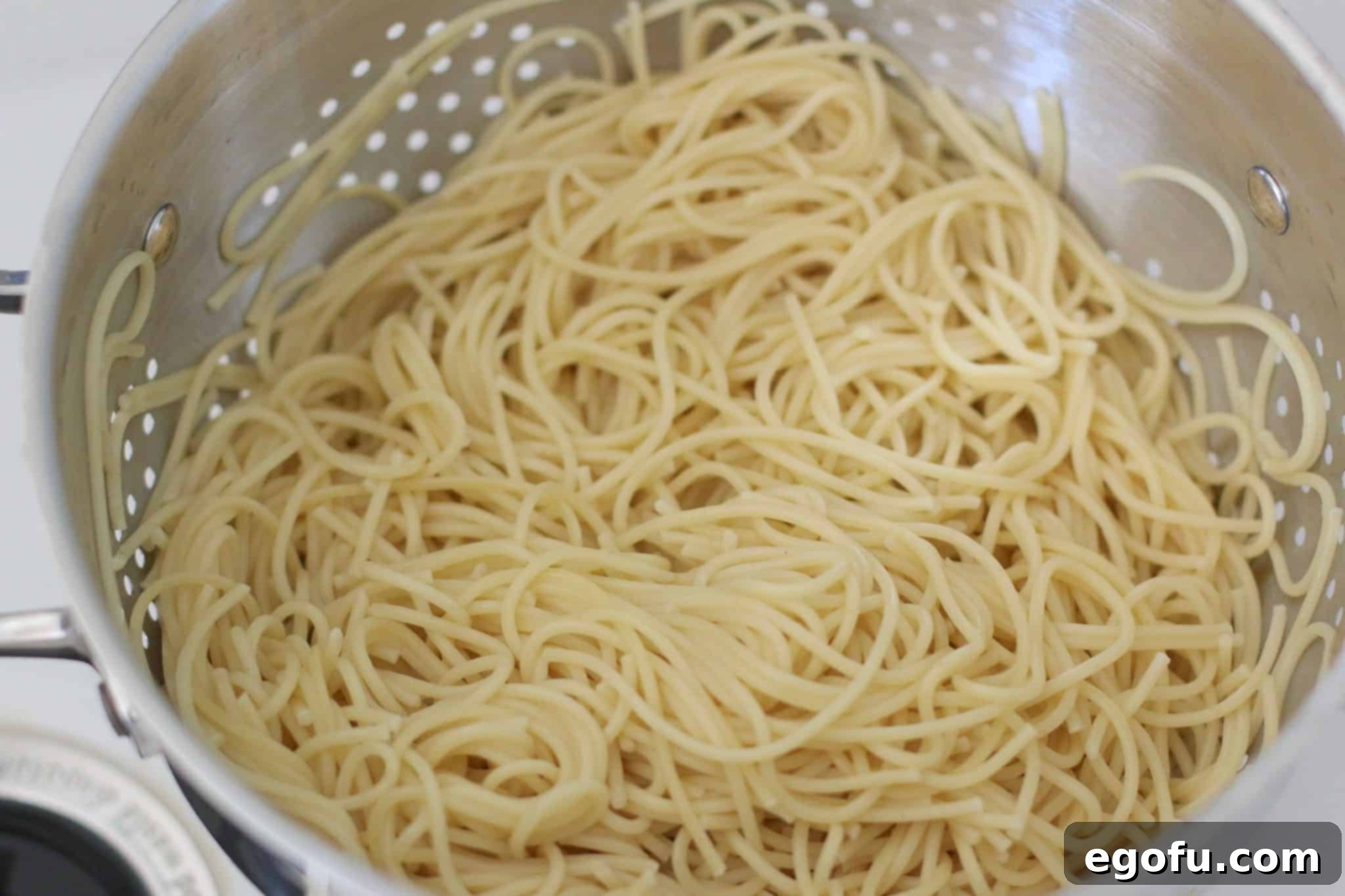 Cooked spaghetti noodles draining thoroughly in a metal strainer, ready to be cooled and added to the salad.