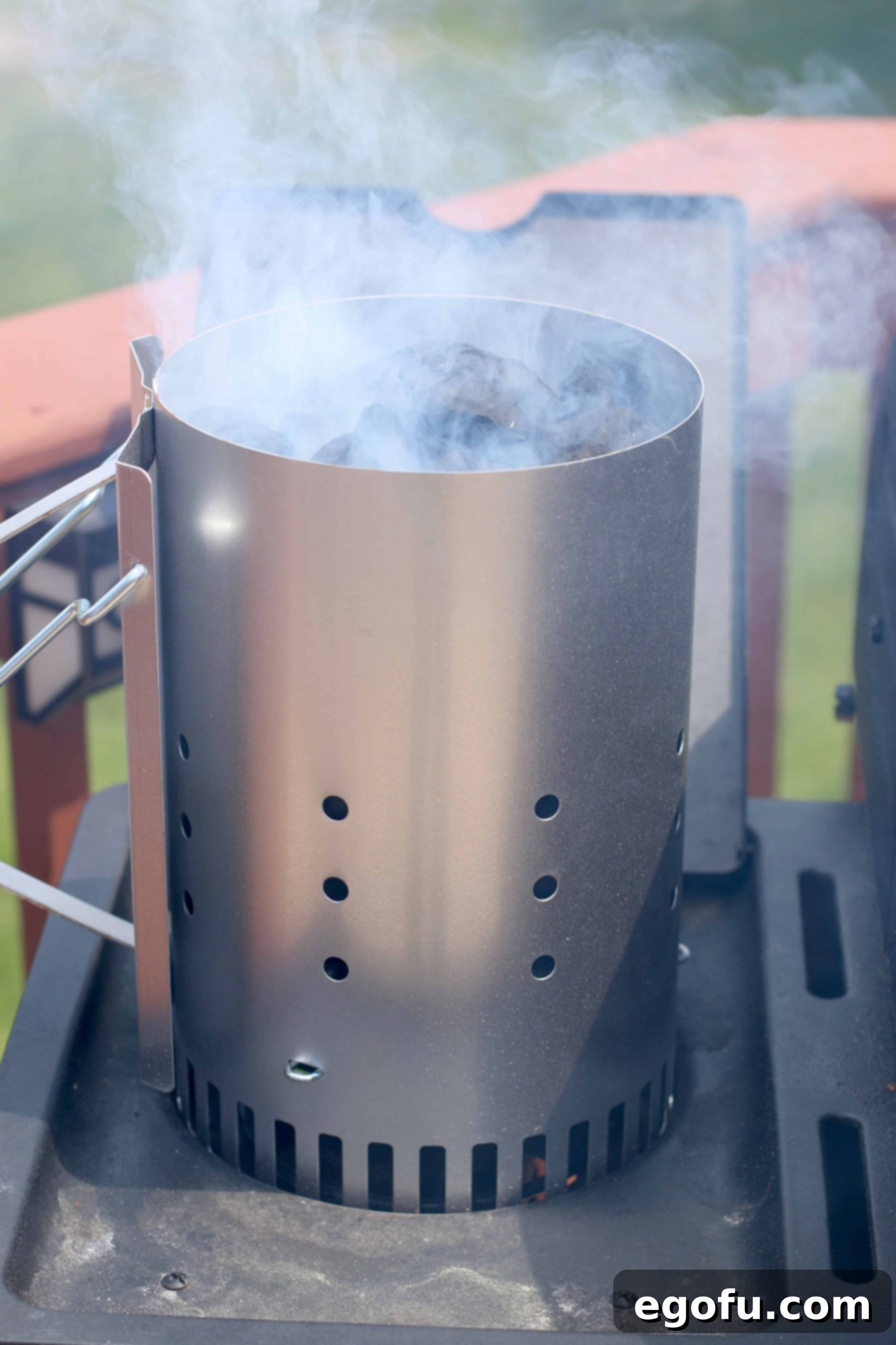 A chimney starter with glowing charcoal briquets producing smoke, indicating they are almost ready for grilling.