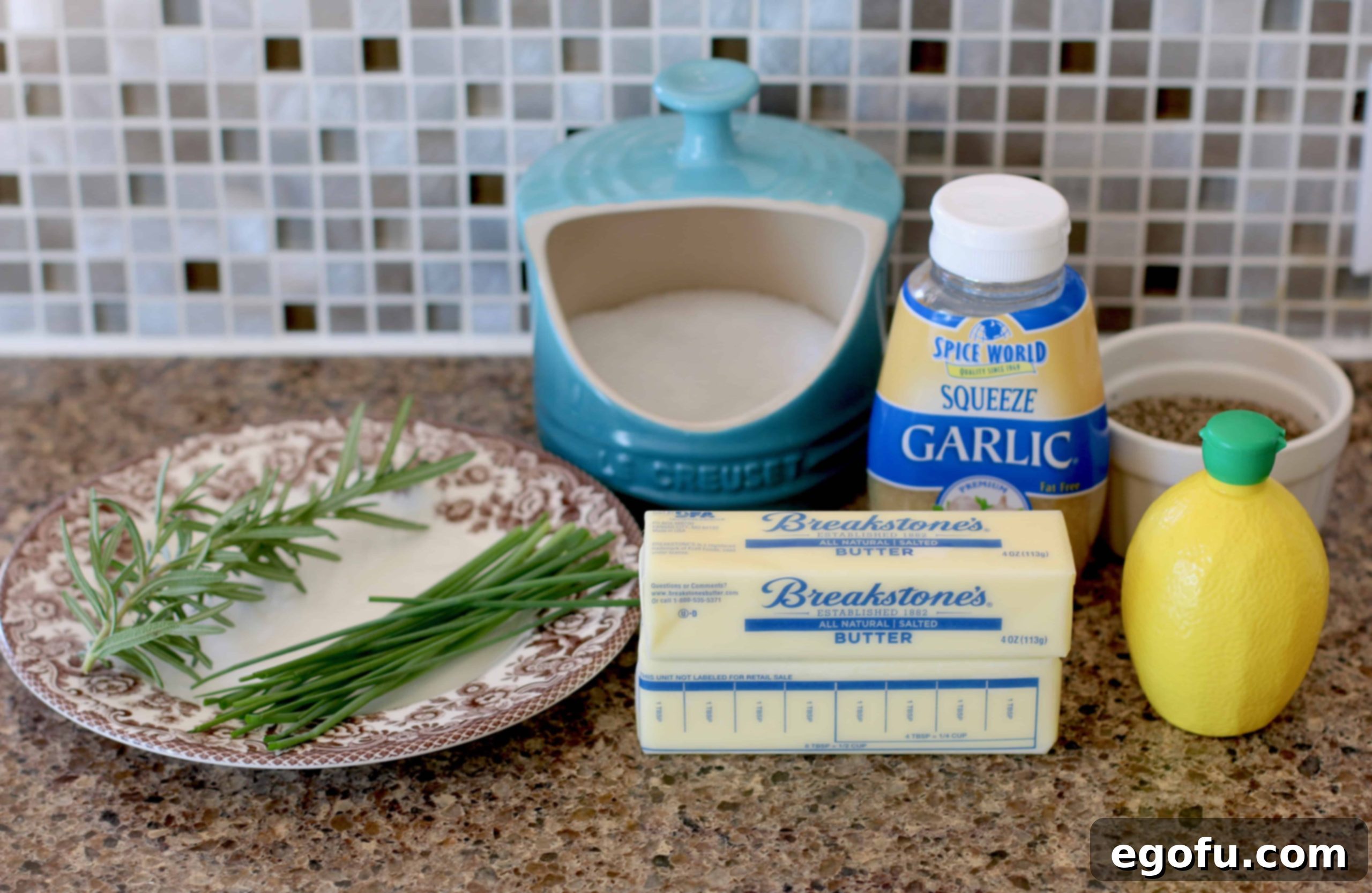The ingredients for steakhouse butter laid out: softened butter, fresh chives, fresh rosemary, minced garlic, lemon juice, kosher salt, and black pepper.