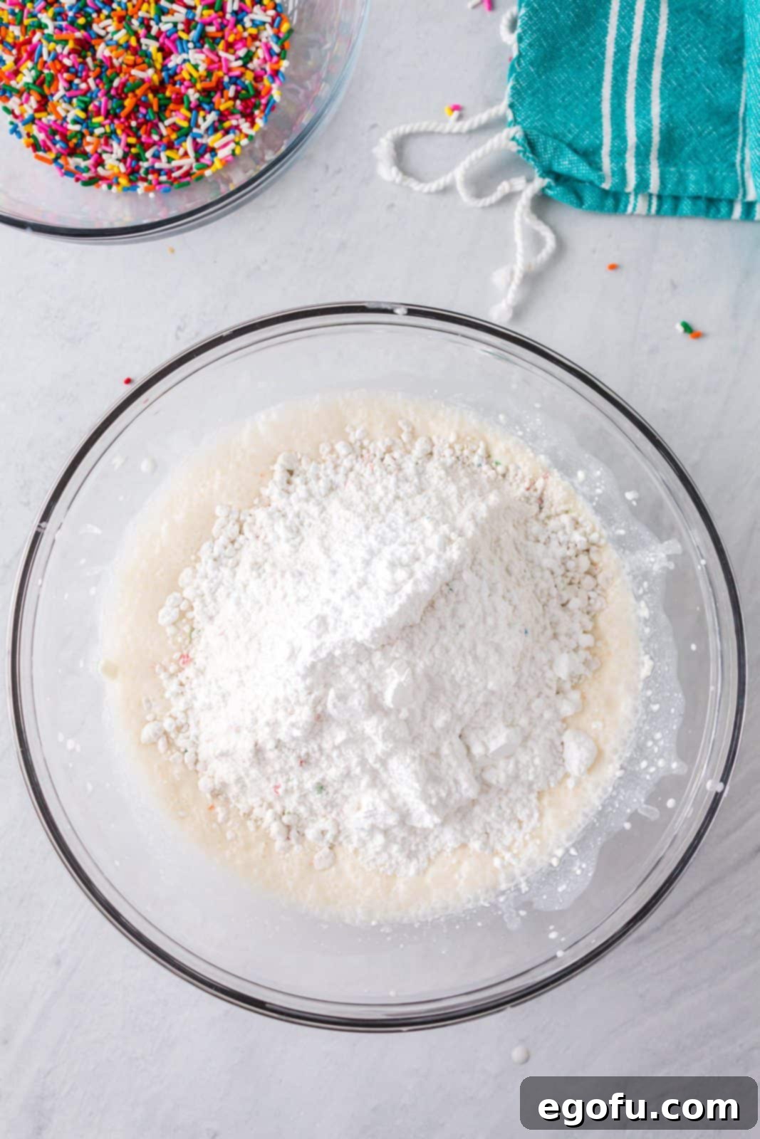 Confetti Cake Bliss 6 Dry cake mix and powdered sugar are being added to the creamy mixture of cream cheese, milk, and vanilla extract in a mixing bowl.