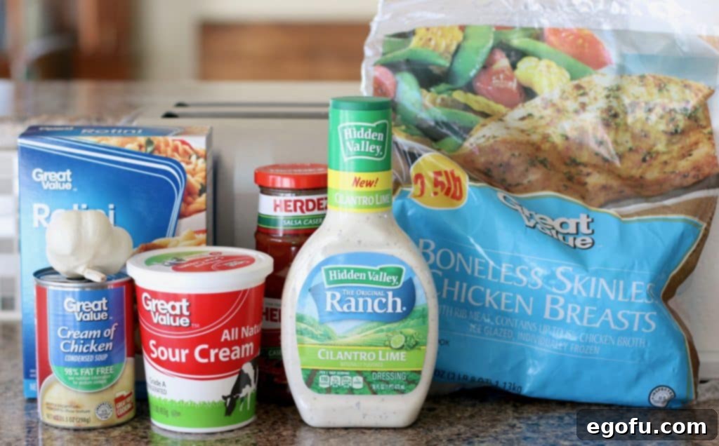 Ingredients for Crock Pot Salsa Ranch Chicken Pasta shown: frozen chicken breasts, ranch dressing, sour cream, rotini pasta, and salsa on a wooden surface.