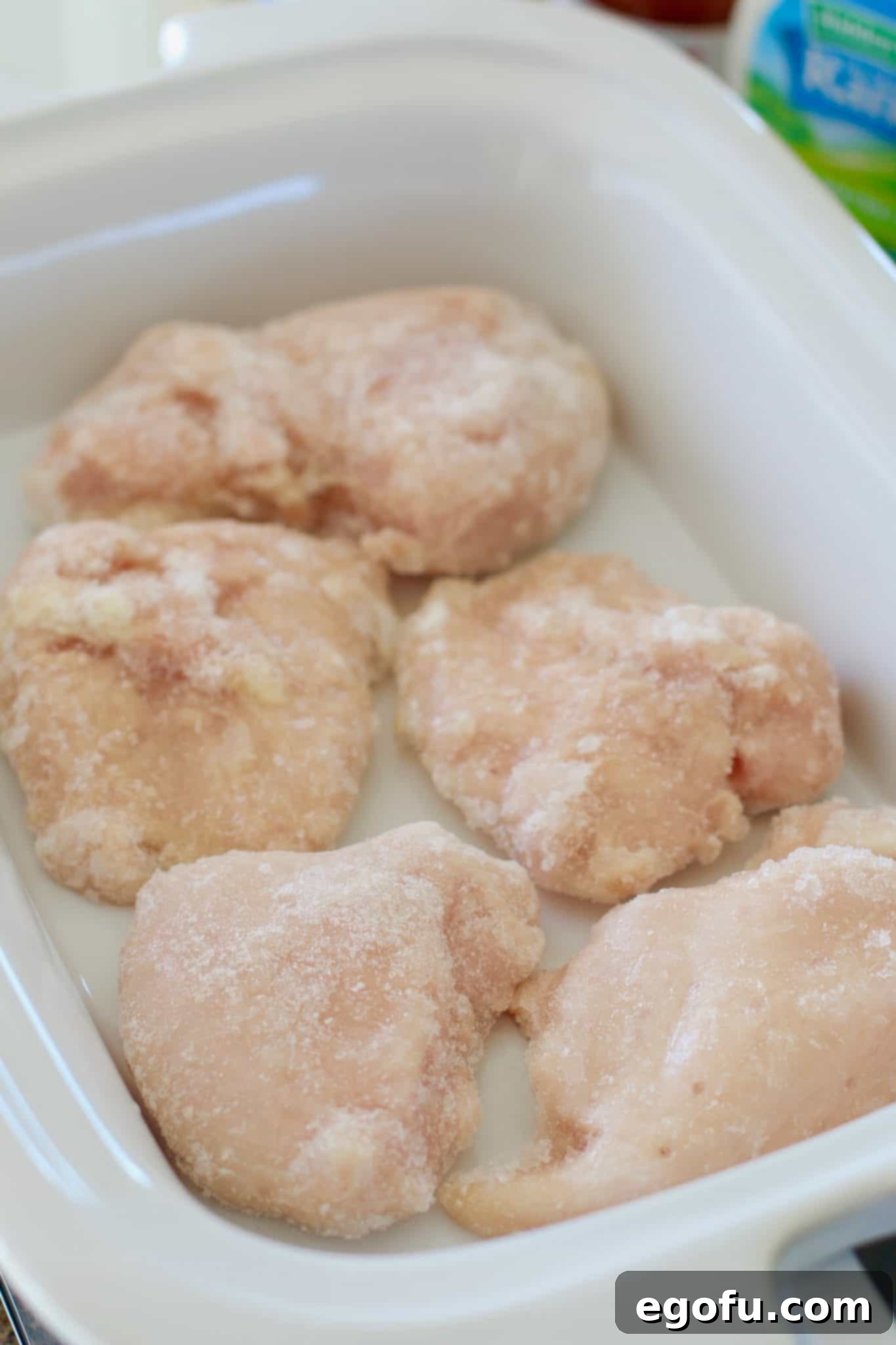 Five frozen chicken breasts arranged in the bottom of a white casserole slow cooker.
