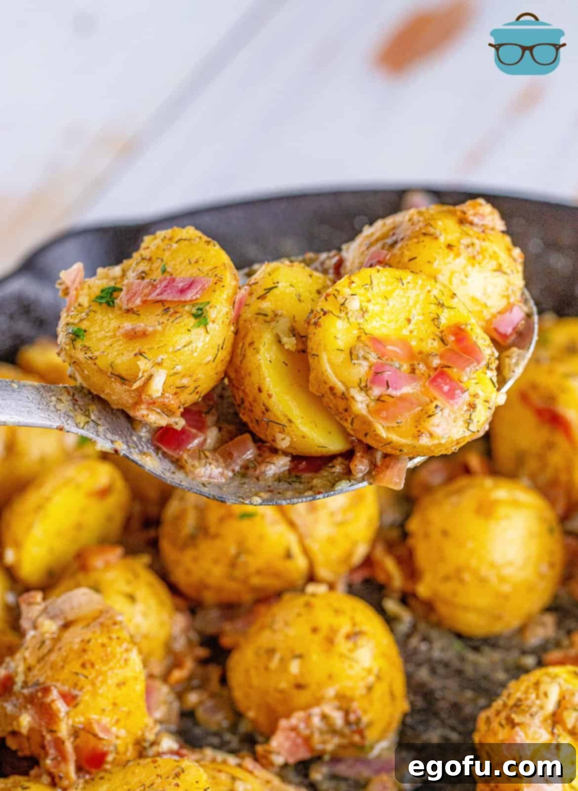 A large serving spoon lifting a portion of warm bacon potato salad from a skillet, showcasing its appealing texture and ingredients.