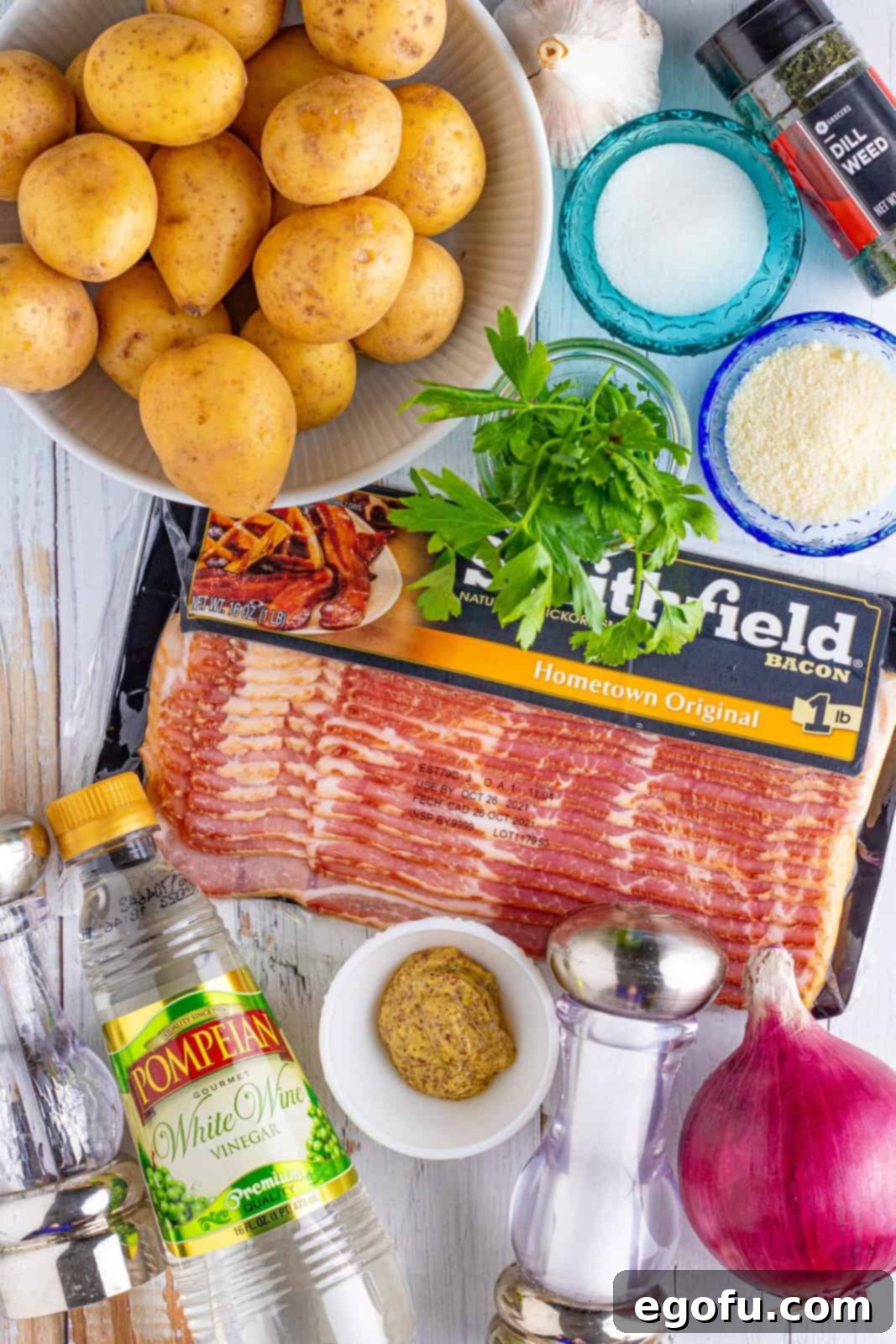 Individual ingredients for warm potato salad laid out: baby gold potatoes, uncooked bacon, red onion, grainy mustard, white wine vinegar, fresh parsley, dried dill, minced garlic, and grated Parmesan cheese.