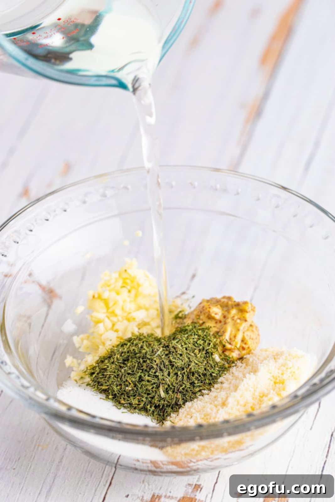 White wine vinegar being poured into a bowl containing sugar, coarse grain mustard, minced garlic, dried dill, and Parmesan cheese, in preparation for whisking the vinaigrette.