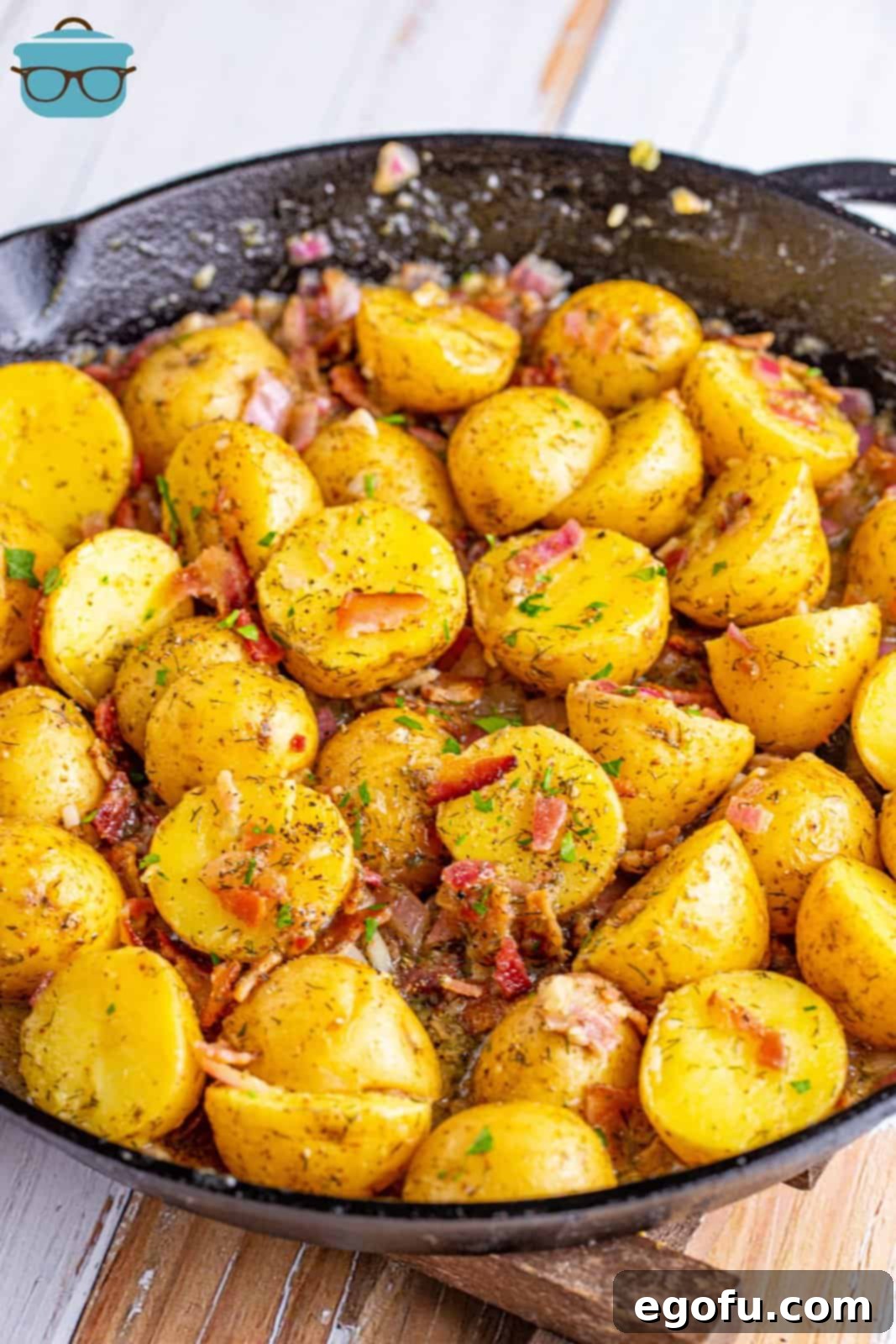 A large cast iron skillet filled with fully cooked warm bacon potato salad, garnished with a sprinkle of fresh chopped parsley on top.