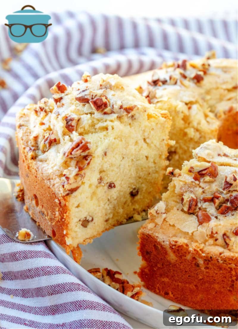 Heirloom Southern Pecan Pound Cake 2 A generous slice of moist Southern Pecan Pound Cake being carefully lifted from a beautifully baked whole bundt cake, presented on a pristine white platter alongside a charming striped kitchen towel.