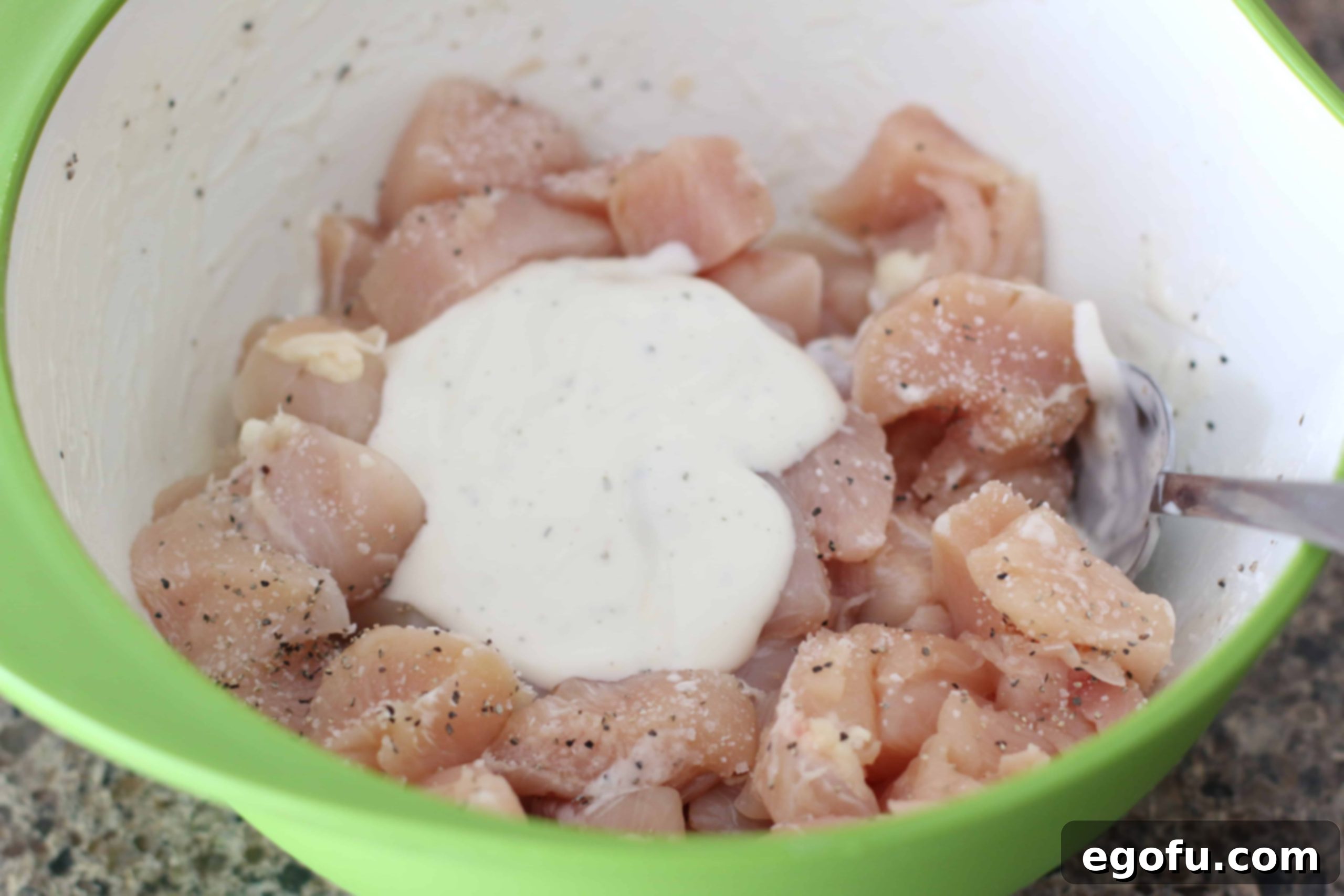 Diced chicken breasts, ranch dressing, salt, and pepper in a bowl with a spoon for marinating.