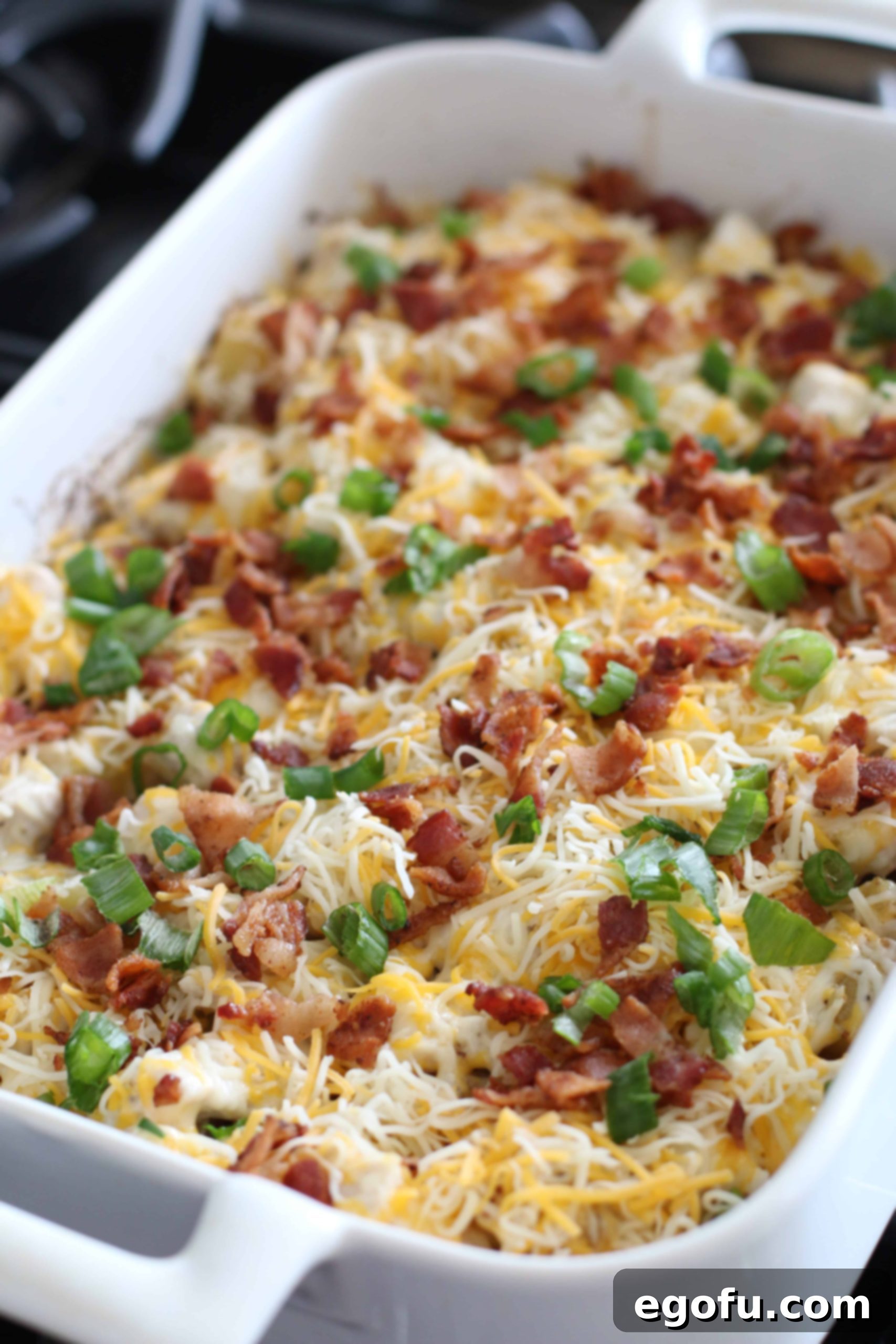 Shredded cheese, bacon, and green onion being layered over diced potatoes and chicken in a white baking dish.