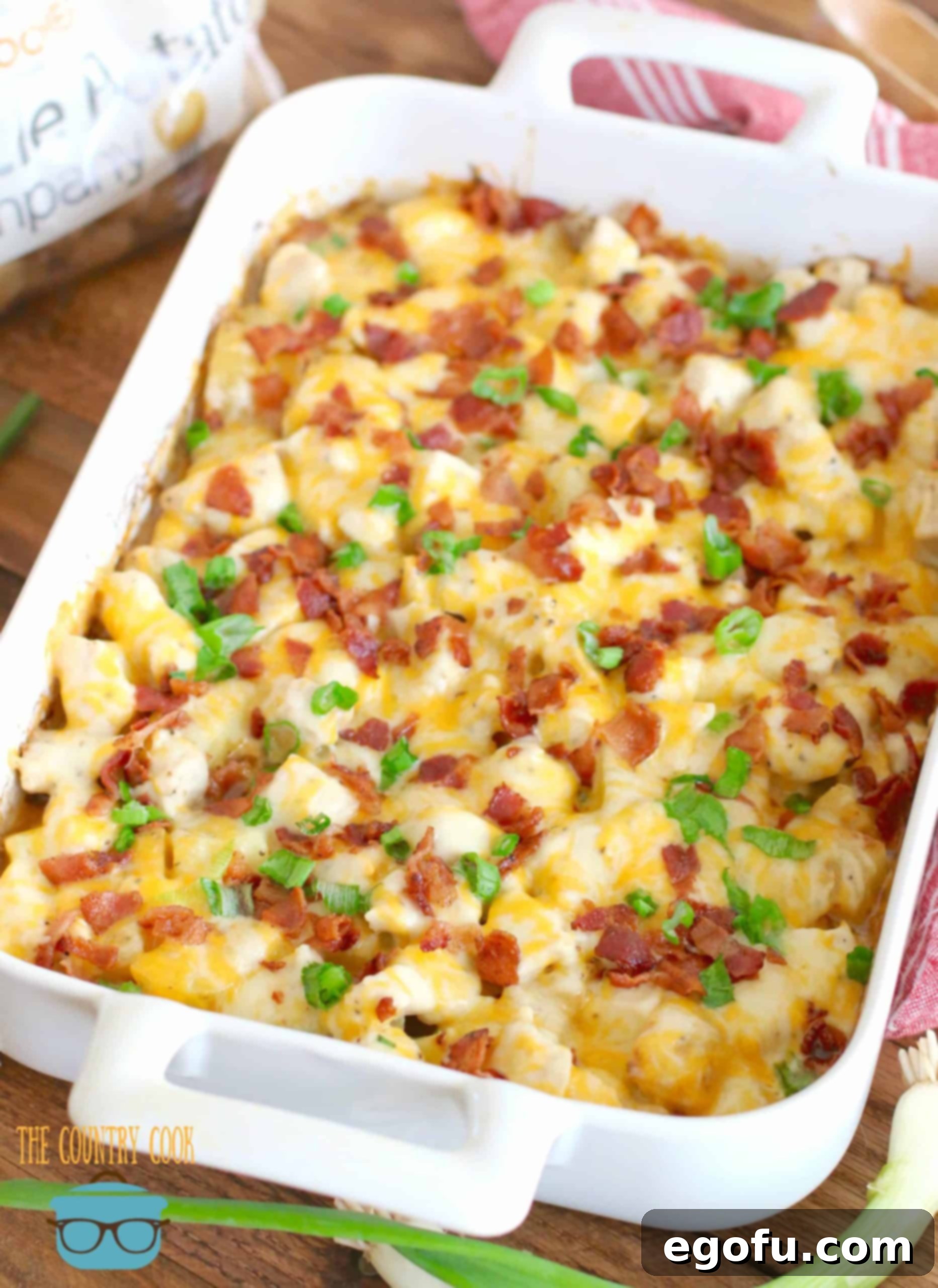 Finished Loaded Potato Ranch Chicken Casserole recipe shown in a white casserole dish on a wooden board, topped with melted cheese, bacon, and green onions.