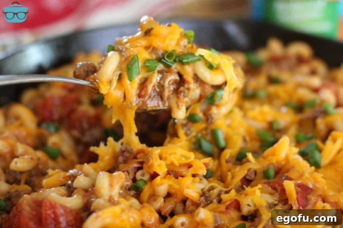 A spoon scooping a generous portion of cheesy tomato macaroni from a rustic cast iron skillet, highlighting the rich texture and melty cheese.