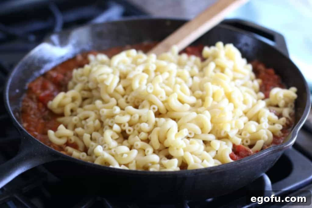 Cooked macaroni noodles being gently folded into the luscious tomato and ground beef sauce in the skillet, ready to absorb all the delicious flavors.
