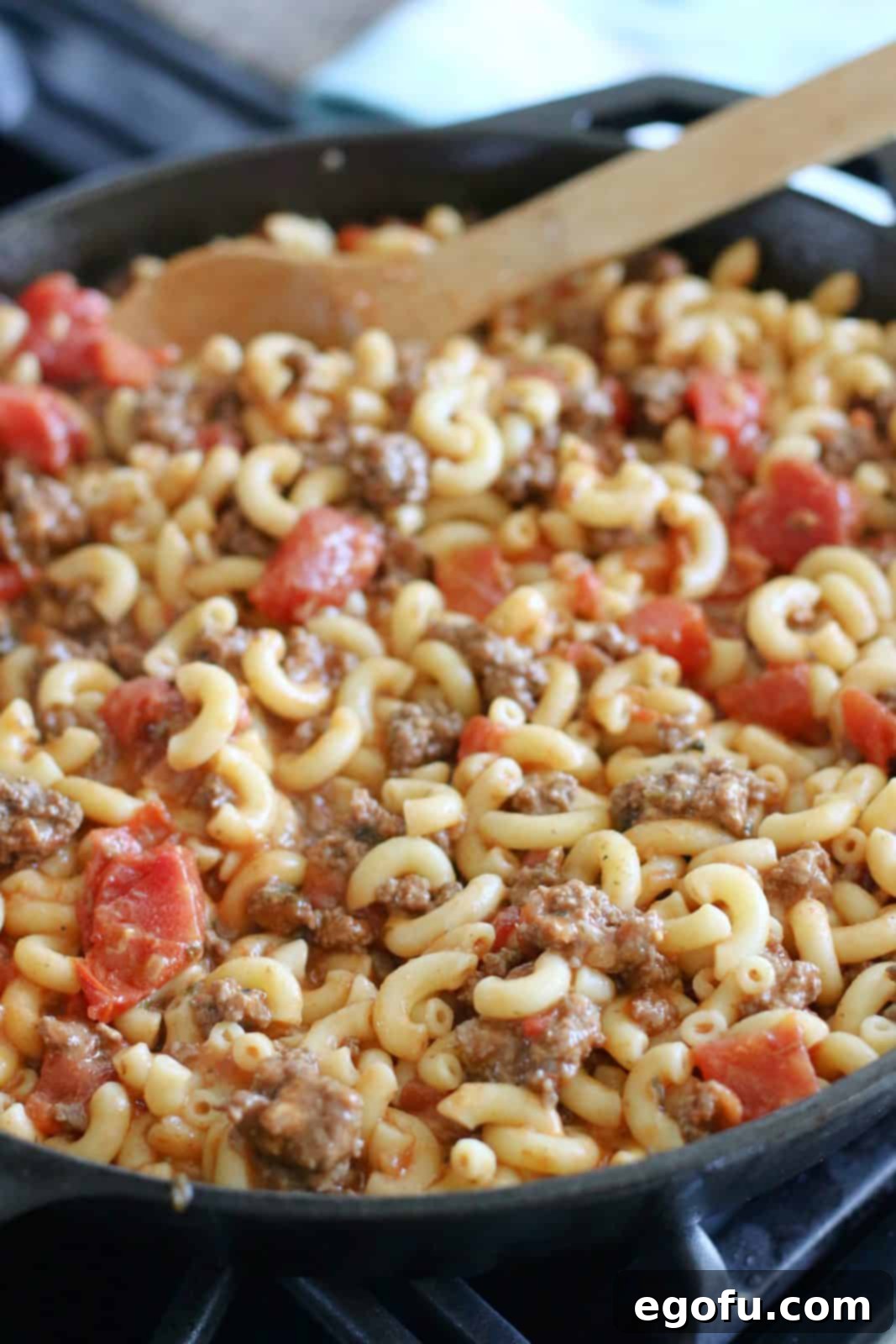 The fully assembled macaroni skillet, a hearty blend of pasta, ground beef, and tomato, resting in a large black cast iron pan, perfectly warmed and ready for the final touch of cheese.