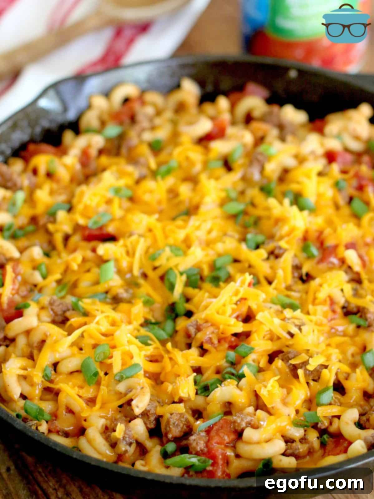 A glorious close-up of shredded cheddar cheese melting beautifully over the top of the macaroni, ground beef, and tomatoes in a cast iron skillet, promising a rich and gooey finish.