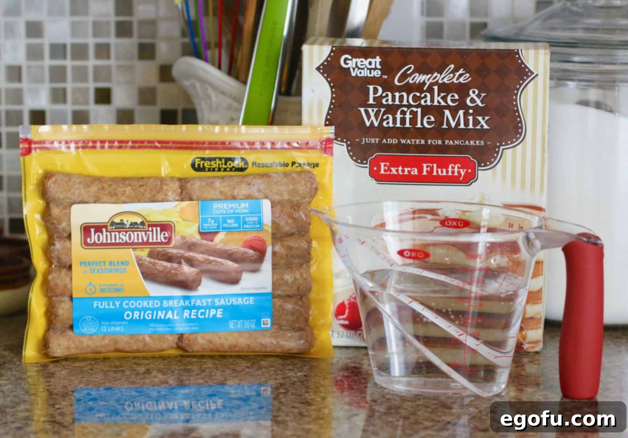 Key ingredients for pancake sausage muffins laid out: a bag of pancake mix, a package of cooked sausages, a glass of water, and a small bottle of maple extract.