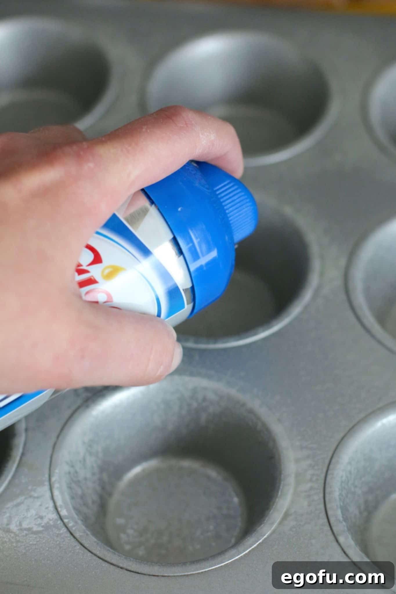 A hand is shown spraying nonstick cooking spray into the cups of a muffin pan, preparing it for baking.