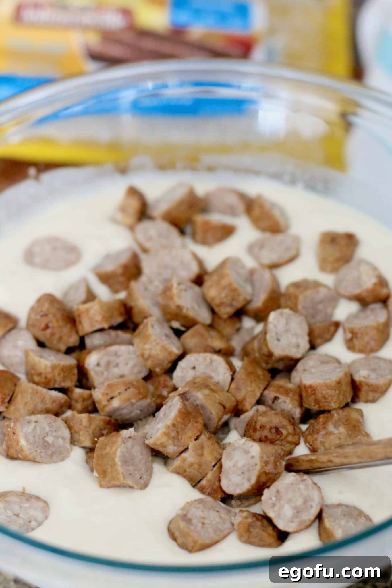 Sliced cooked breakfast sausages are shown resting on top of the prepared pancake batter in a mixing bowl, ready to be folded in.