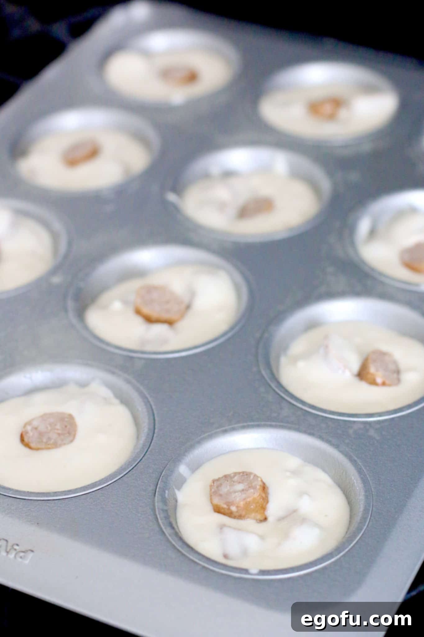 Muffin tin cups filled about three-quarters with pancake batter and visible slices of sausage distributed within, ready for baking.