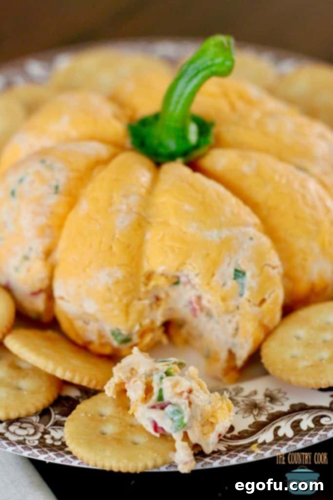 The Great Pumpkin Cheese Ball 3 Vegetable Cream Cheese Cheeseball covered in shredded cheddat cheese and shaped into a pumpkin. A piece shown removed and served on a Ritz cracerk.