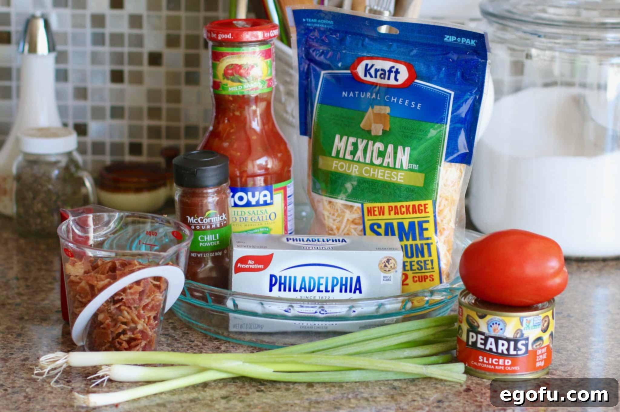 Hot Cheesy Bacon Dip 3 Assorted ingredients laid out: cooked bacon pieces, diced tomato, sliced black olives, softened cream cheese, shredded cheese, salsa, and green onions, chili powder.