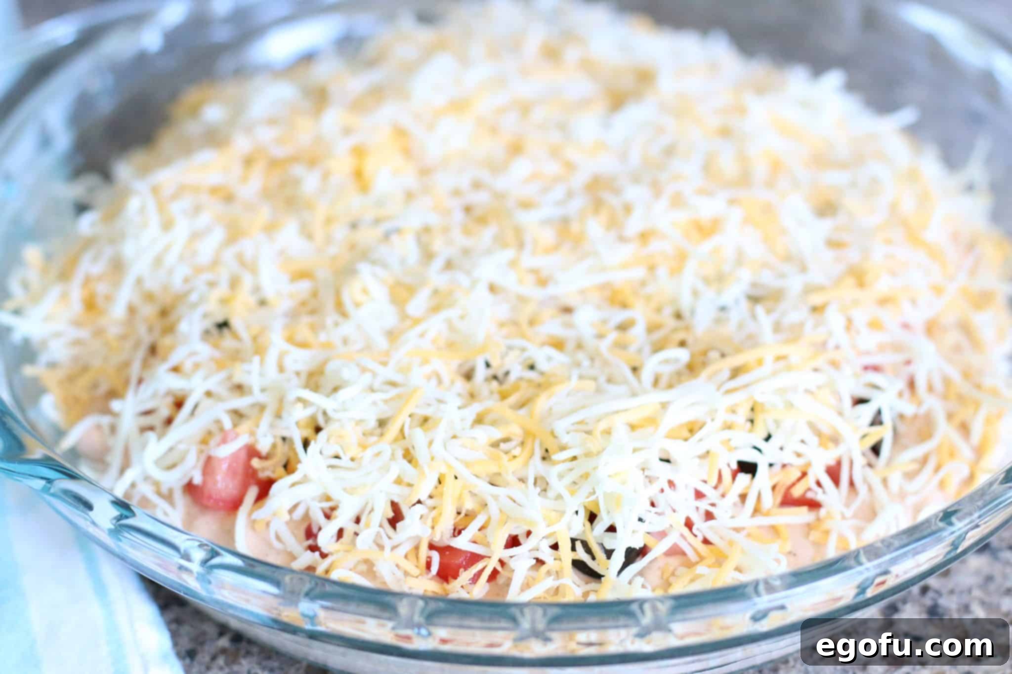 Hot Cheesy Bacon Dip 7 Shredded cheddar cheese generously covering the dip mixture with olives and tomatoes in a round baking dish.