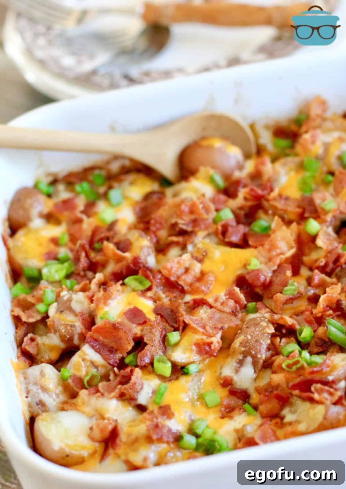 A wooden spoon inserted into a white dish containing cooked baby potatoes with cheese and bacon.
