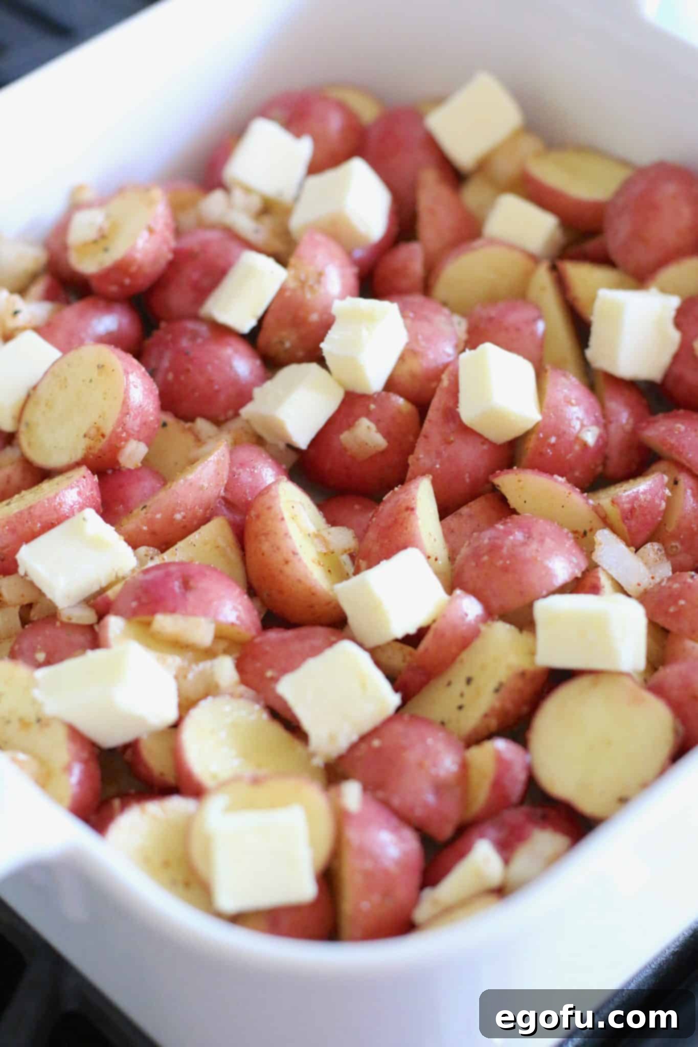 Sliced baby potatoes with seasoning topped with salted butter in a square baking dish.