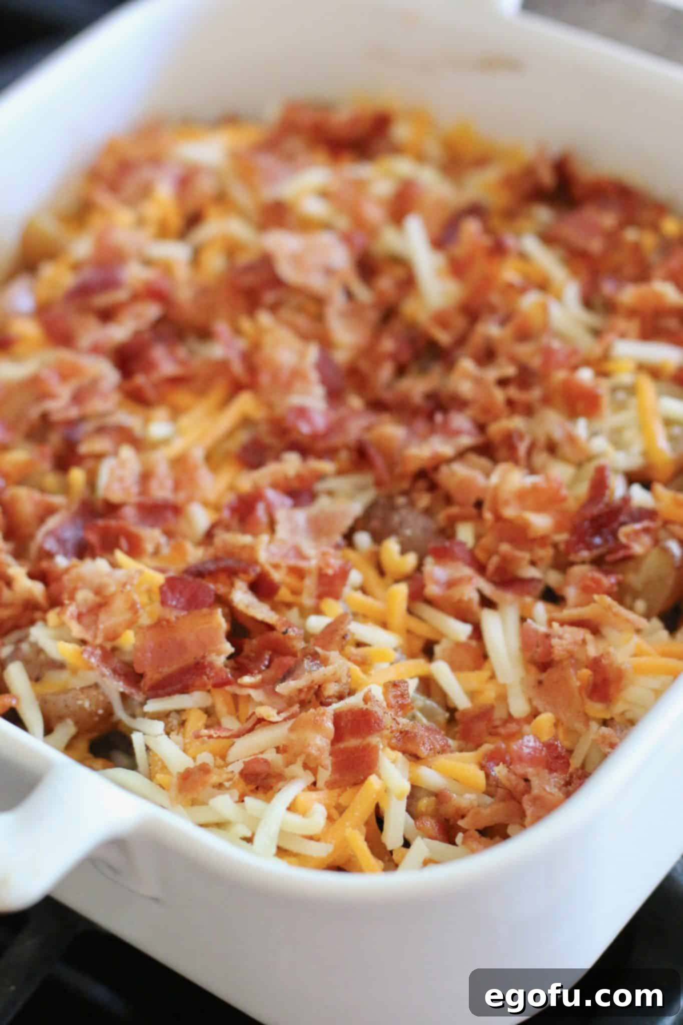 Baked baby potatoes topped with shredded cheddar cheese and cooked and crumbled bacon in a square 8x8 white Revol baking dish.