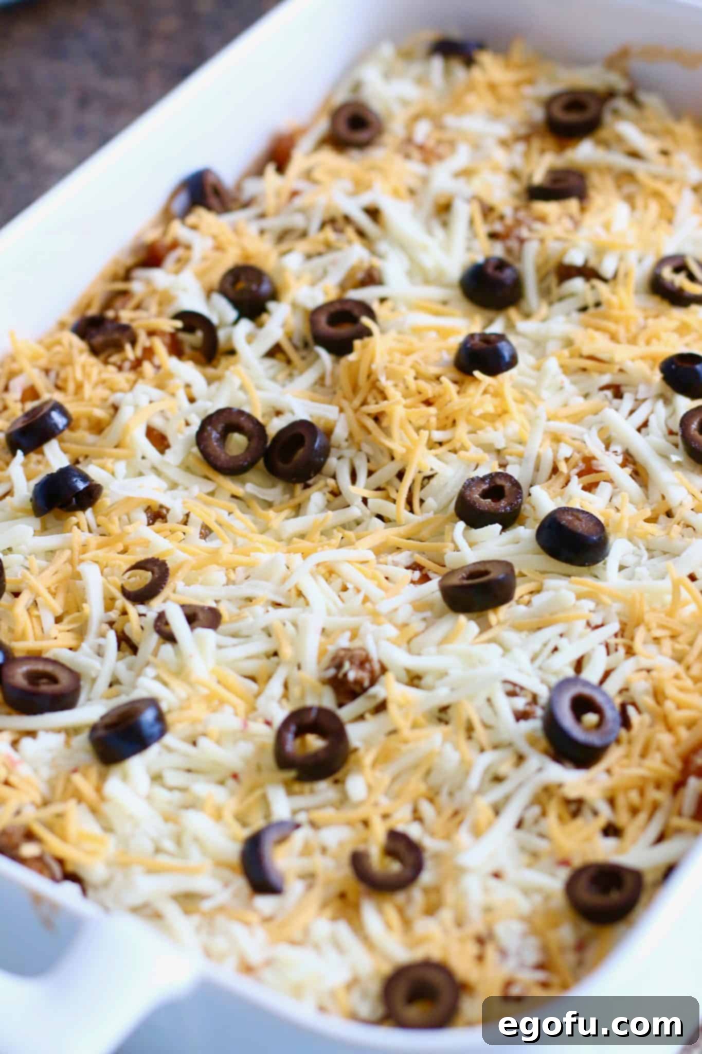 Shredded cheese and sliced olives topping an enchilada casserole, ready for the oven.