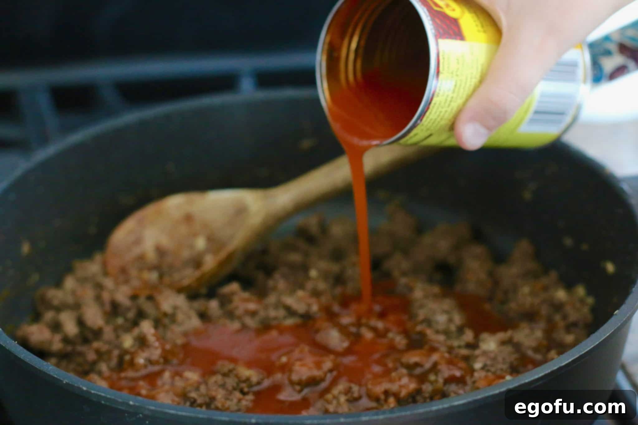 A stream of red enchilada sauce being poured into the ground beef mixture in a skillet, adding moisture and flavor.