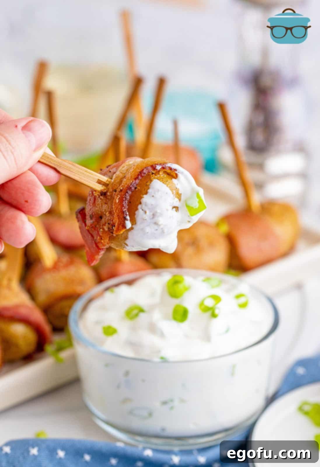 A hand dipping a bacon-wrapped potato into a bowl of sour cream and green onion dip.