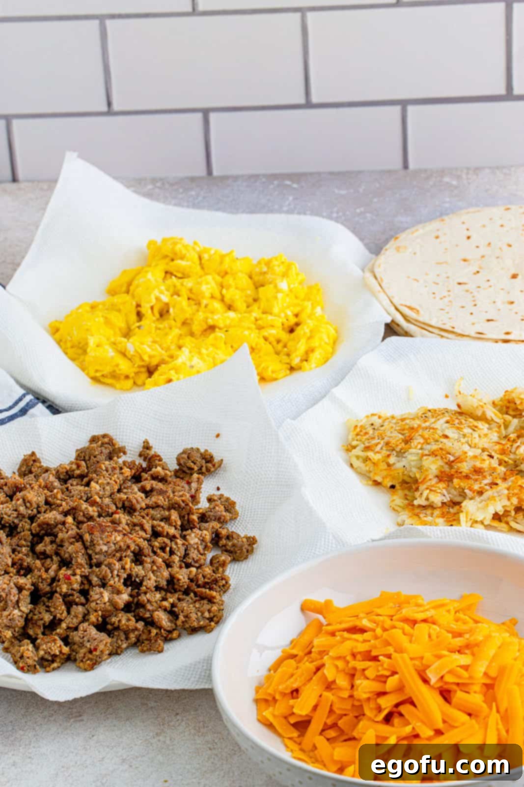 An organized assembly line of all the prepared breakfast burrito ingredients: cooked sausage, scrambled eggs, shredded cheese, and crispy hash browns, ready for wrapping.