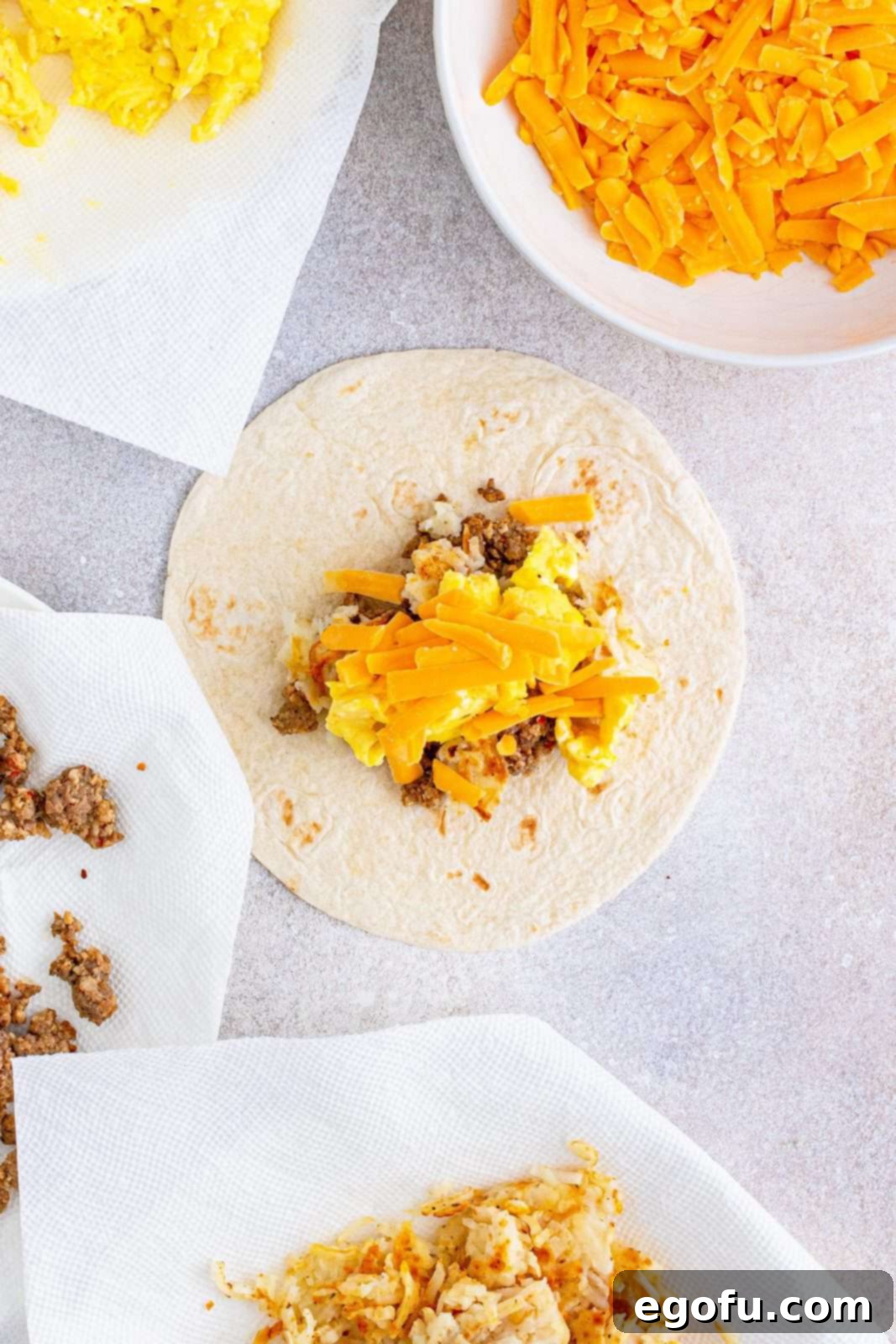 A soft flour tortilla laid flat, with generous portions of scrambled eggs, sausage, shredded cheese, and crispy hash browns neatly arranged down the center, ready for rolling.
