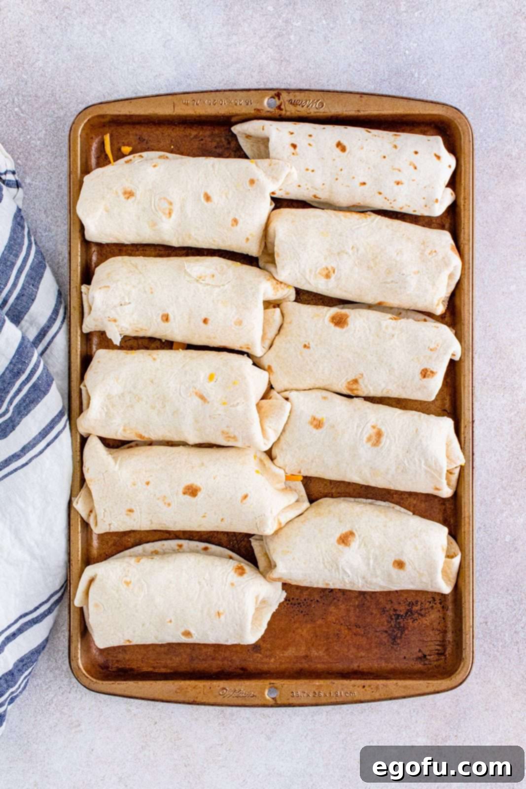 A row of ten perfectly folded breakfast burritos, neatly arranged on a baking tray, ready for wrapping or immediate enjoyment.