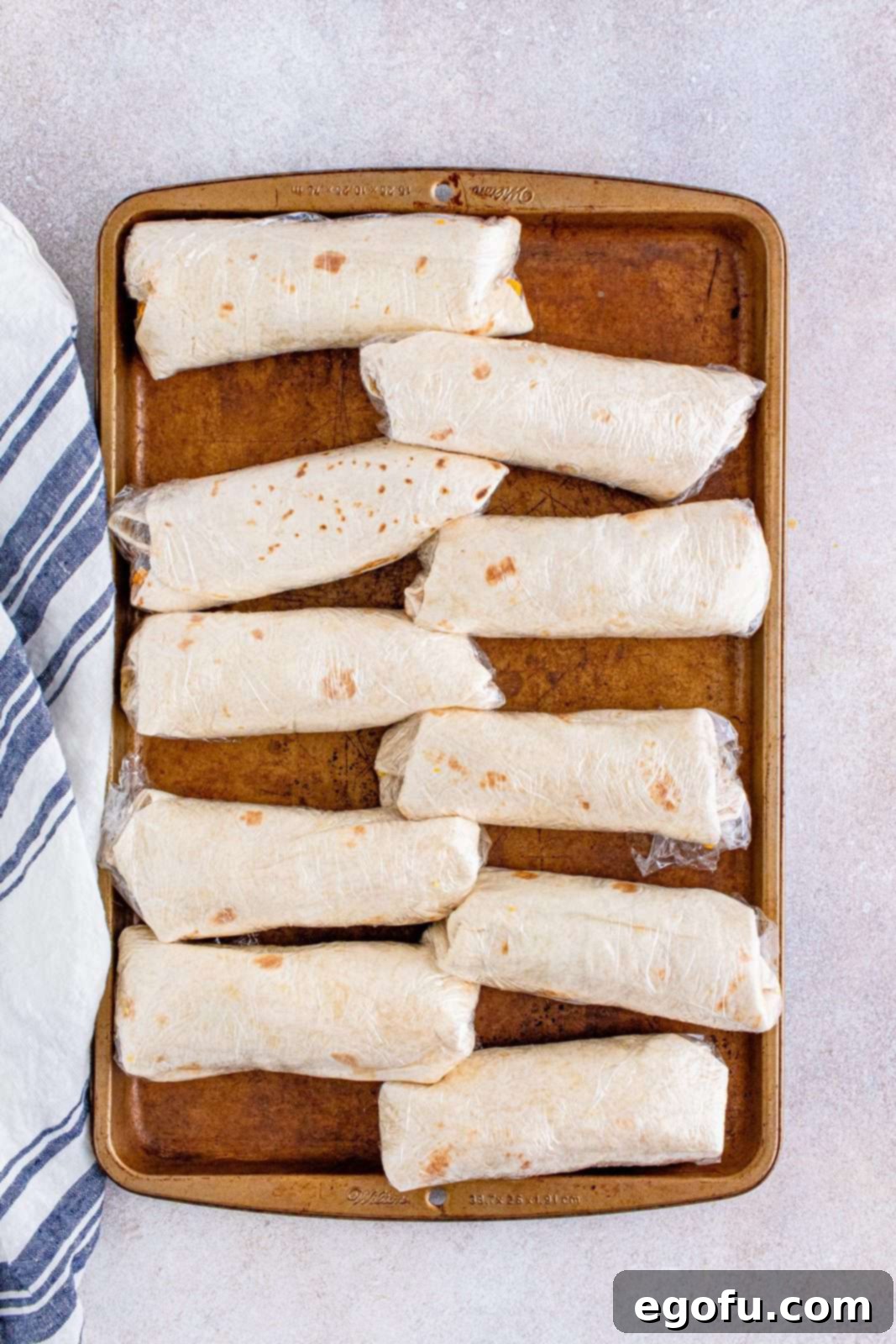 Individual breakfast burritos, each meticulously wrapped in clear plastic wrap, arranged on a baking tray, ready for freezing.