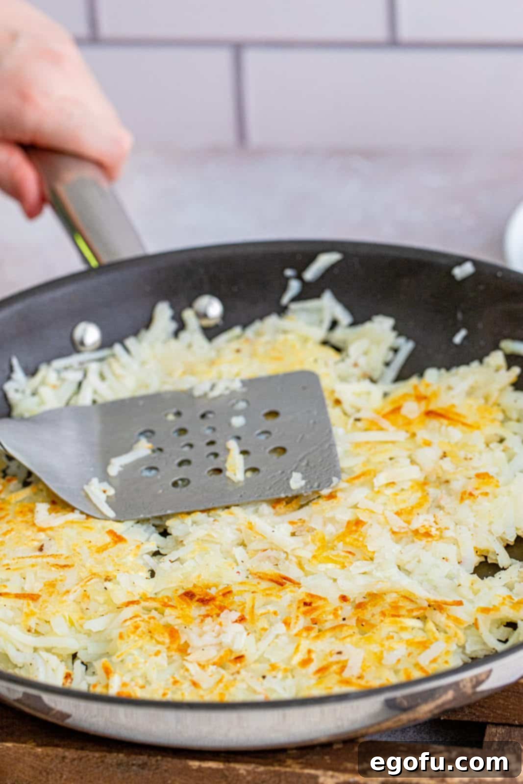 A spatula pressing down shredded hash browns in a large skillet, ensuring they cook in a single layer for maximum crispiness.