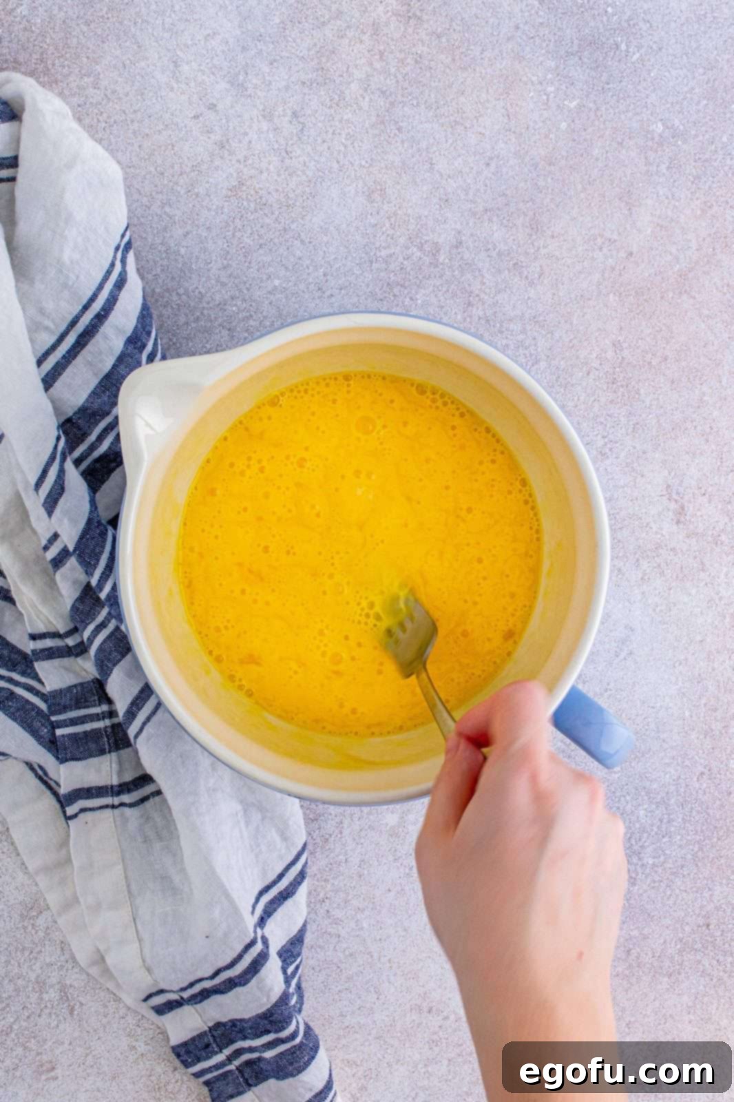 A fork vigorously mixing eggs and water in a white bowl, preparing them for scrambling.
