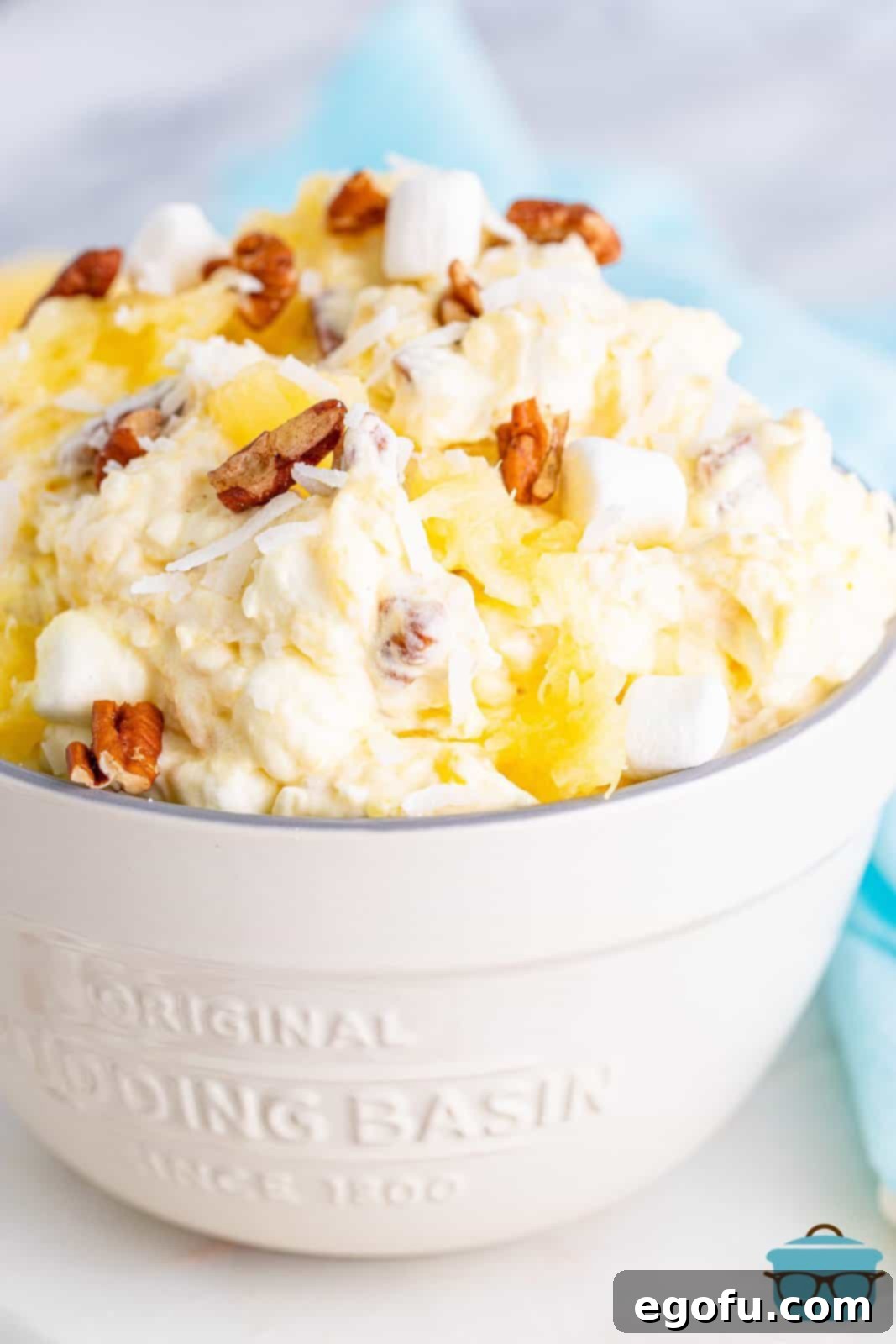 Tropical Cloud Delight 2 A vibrant Pineapple Fluff dessert, generously topped with chopped pecans, presented in a pristine white serving bowl on a rustic white wooden surface. The image captures the dessert's light and airy texture, inviting you to discover its refreshing taste.