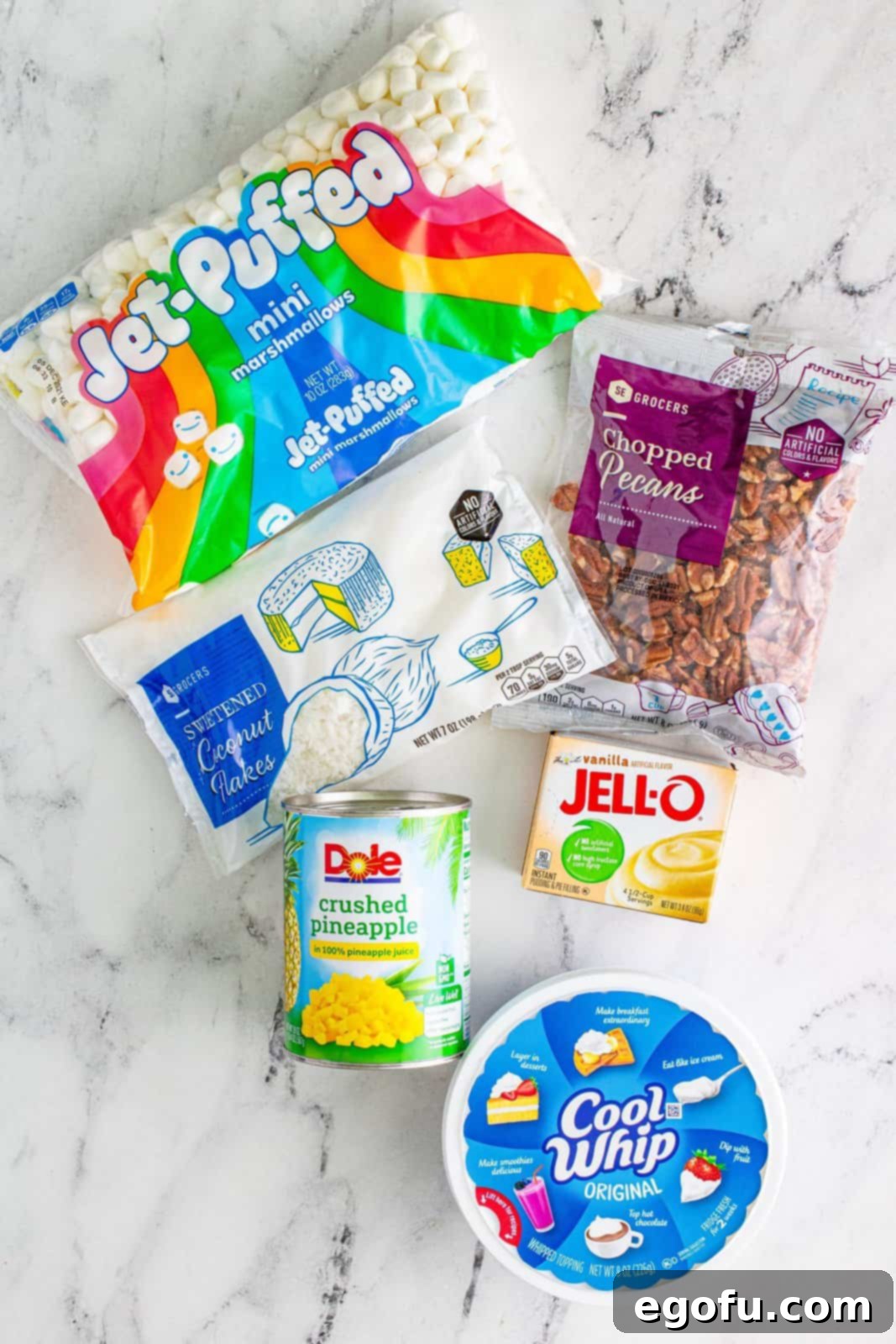 Tropical Cloud Delight 4 All the ingredients for Pineapple Fluff laid out: a can of crushed pineapple, a tub of Cool Whip, a box of instant vanilla pudding mix, a bag of sweetened coconut flakes, a pile of chopped pecans, and a bag of mini marshmallows. Each item is clearly visible, ready for preparation.