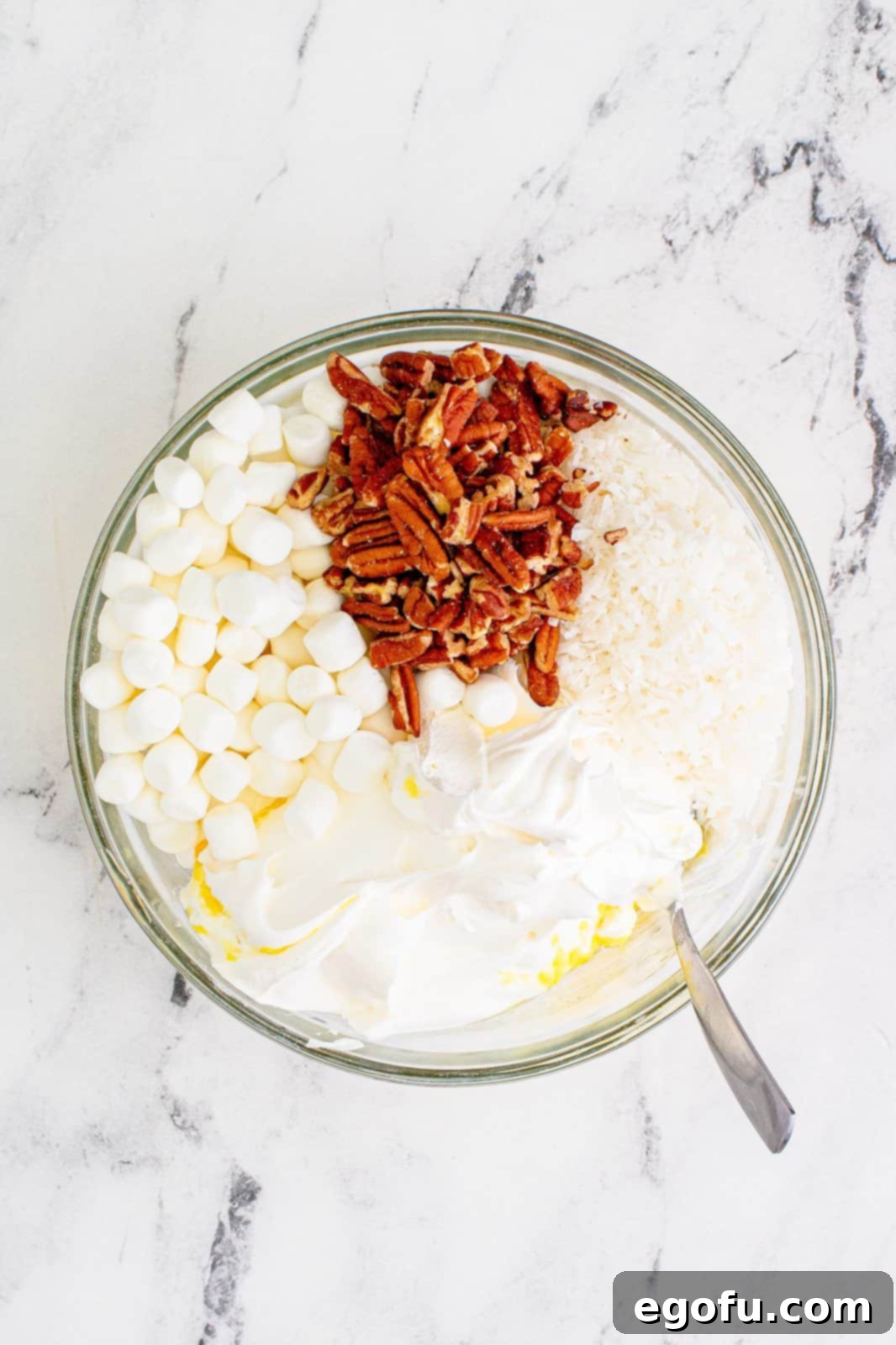 Tropical Cloud Delight 6 Mini marshmallows, chopped pecans, and thawed Cool Whip being gently added to the pineapple and pudding mixture in a clear bowl, showing the next step in assembling the fluff.