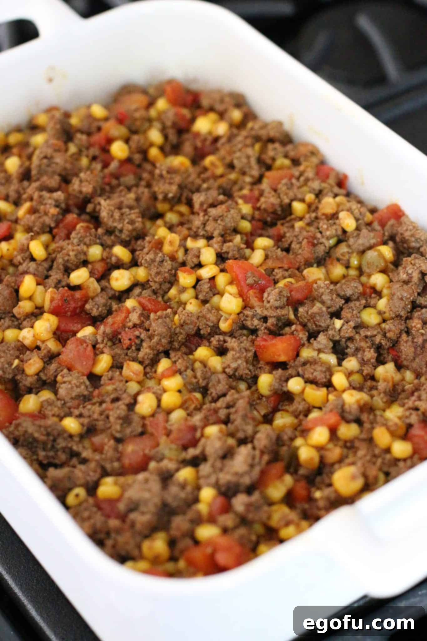 cooked ground beef mixture spread out on top of cornbread in a white baking dish.