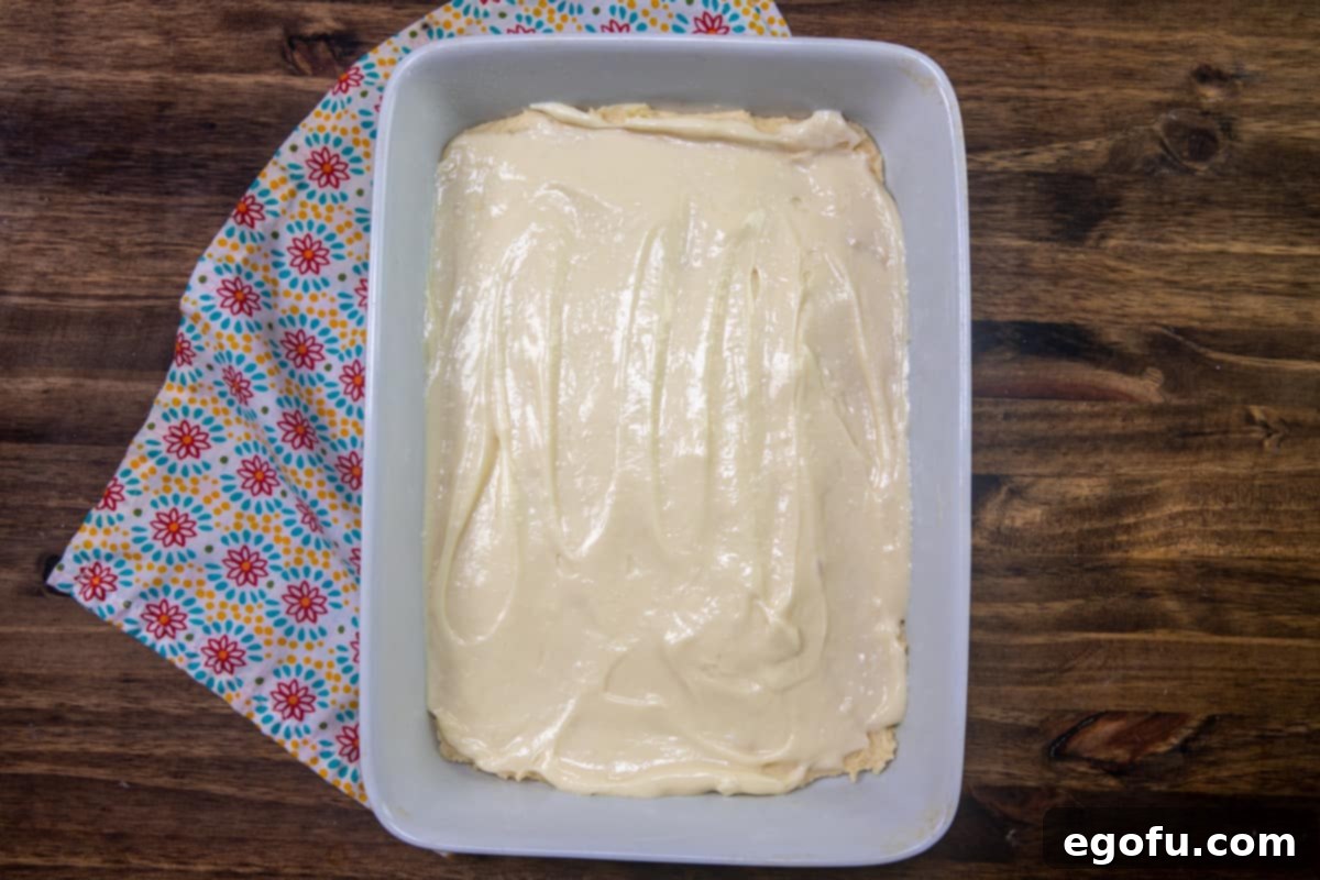 Velvet Peach Cream Bars 10 The creamy cheesecake batter is meticulously spread evenly on top of the first cake batter layer in a large white baking pan, forming a distinct and smooth middle layer for the Peaches and Cream Bars.