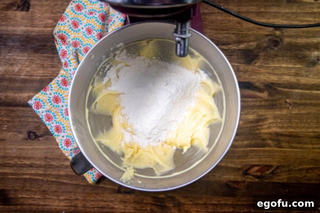 Velvet Peach Cream Bars 7 All-purpose flour is being carefully added to the butter, sugar, and egg mixture in a mixing bowl, preparing for gentle combination to form the bar batter.