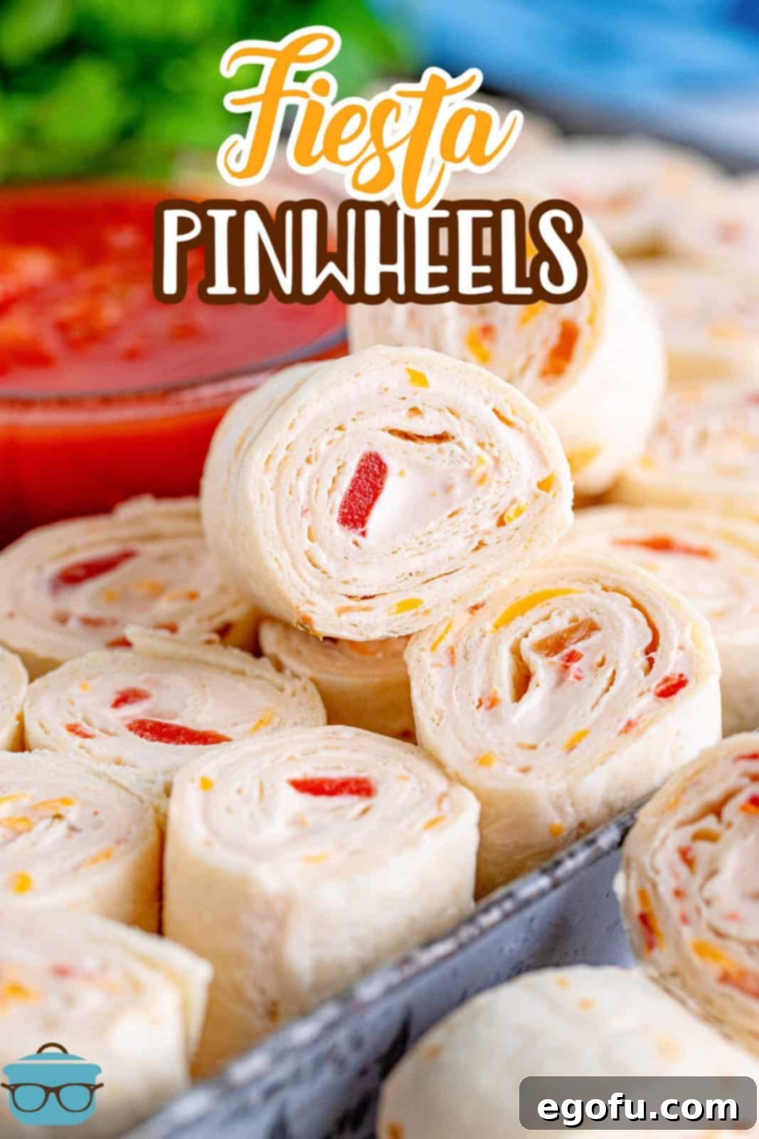 A close-up photograph showcasing a stack of perfectly cut Fiesta Pinwheels, artfully arranged on a rustic metal tray, with a blurred bowl of vibrant red salsa in the background, hinting at their delicious pairing.