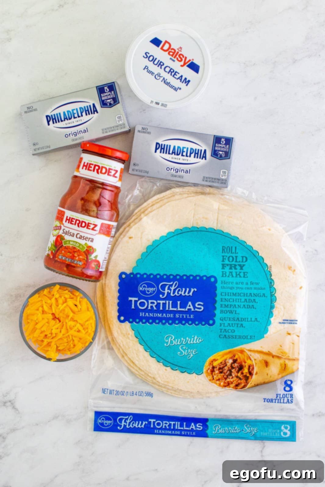 An overhead shot showcasing all the key ingredients for Fiesta Pinwheels neatly arranged on a light surface: soft flour tortillas, a bowl of vibrant red salsa, creamy blocks of cream cheese, a tub of sour cream, and a pile of golden shredded cheddar cheese.