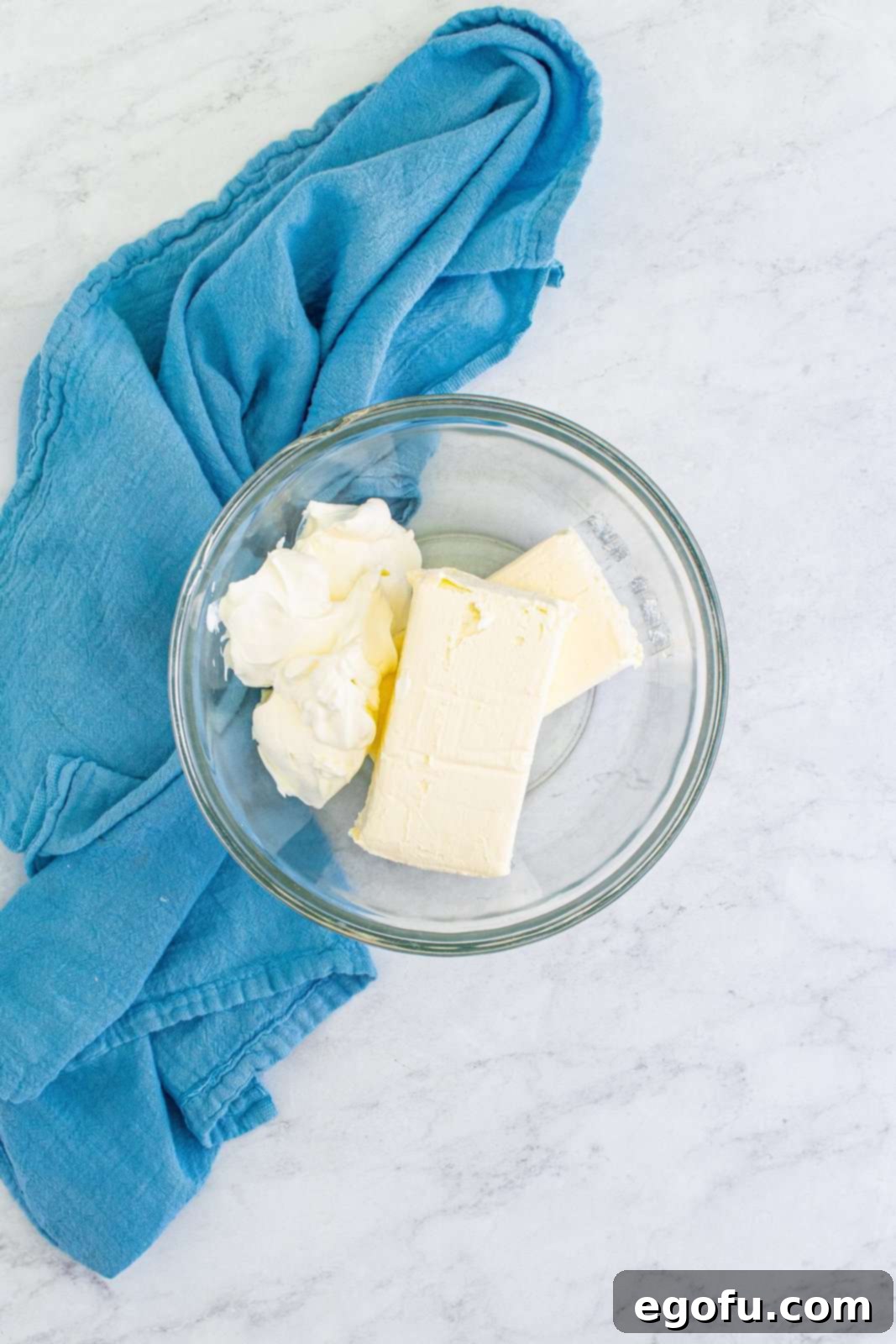 A clear glass bowl contains a smooth, light-colored mixture of cream cheese and sour cream, expertly blended to form a creamy, inviting base, ready for the next ingredients.