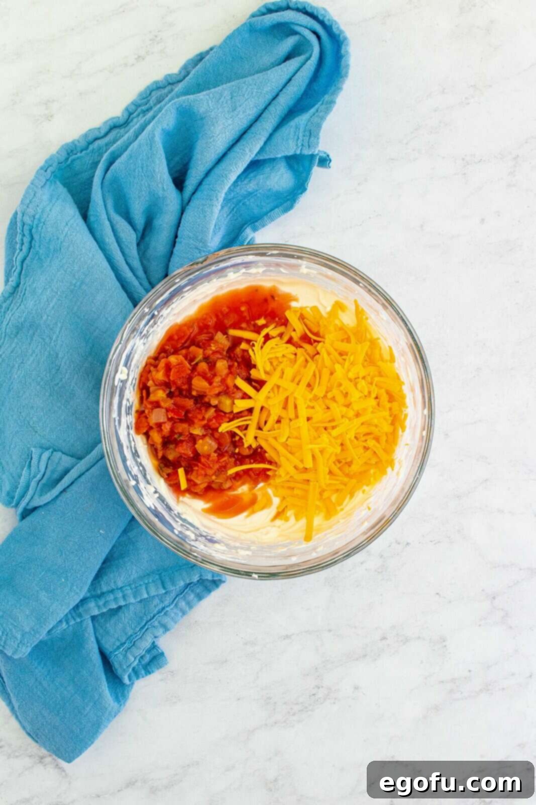 Inside a clear glass bowl, a rich, colorful mixture of cream cheese, sour cream, chunky red salsa, and golden shredded cheddar cheese is blended, showcasing its readiness to become a delicious filling.
